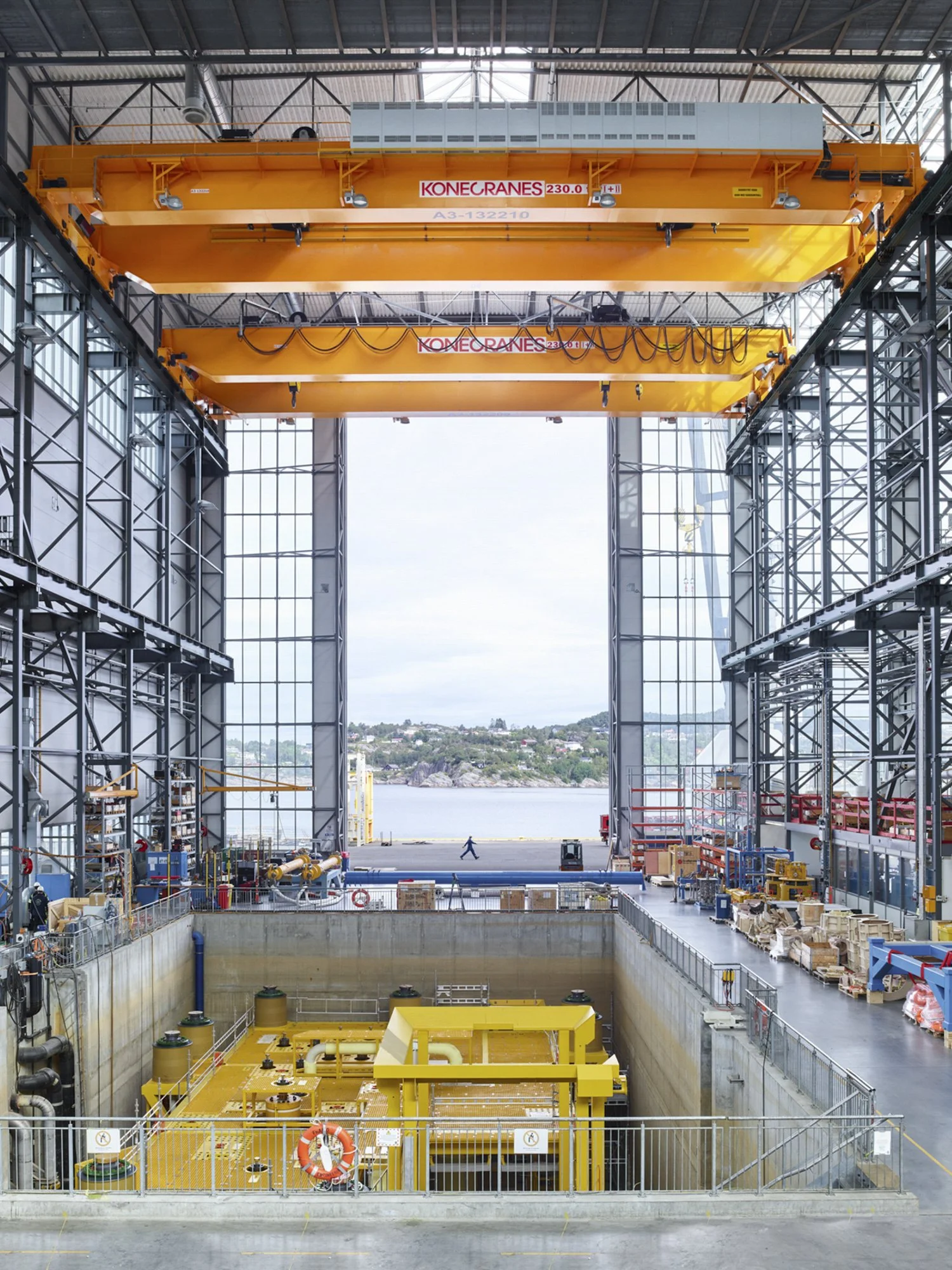 Inside an industrial building featuring two large yellow overhead cranes labeled 'KONECRANES' with max capacities of 230 tons, overlooking a view of water and a hillside with buildings outside.