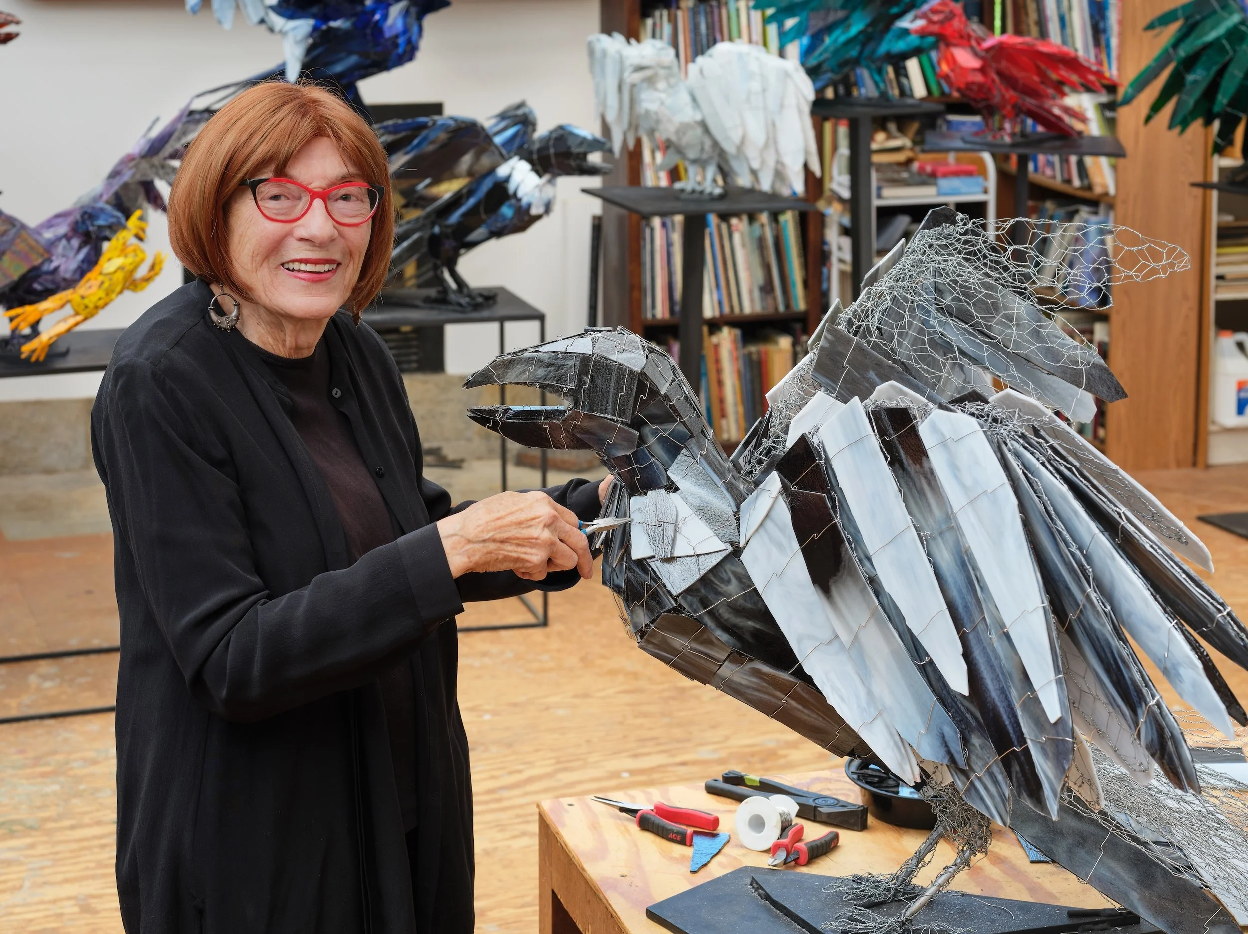 Portrait of Joan Danziger standing in her studio beside a metal and glass Raven sculpture.