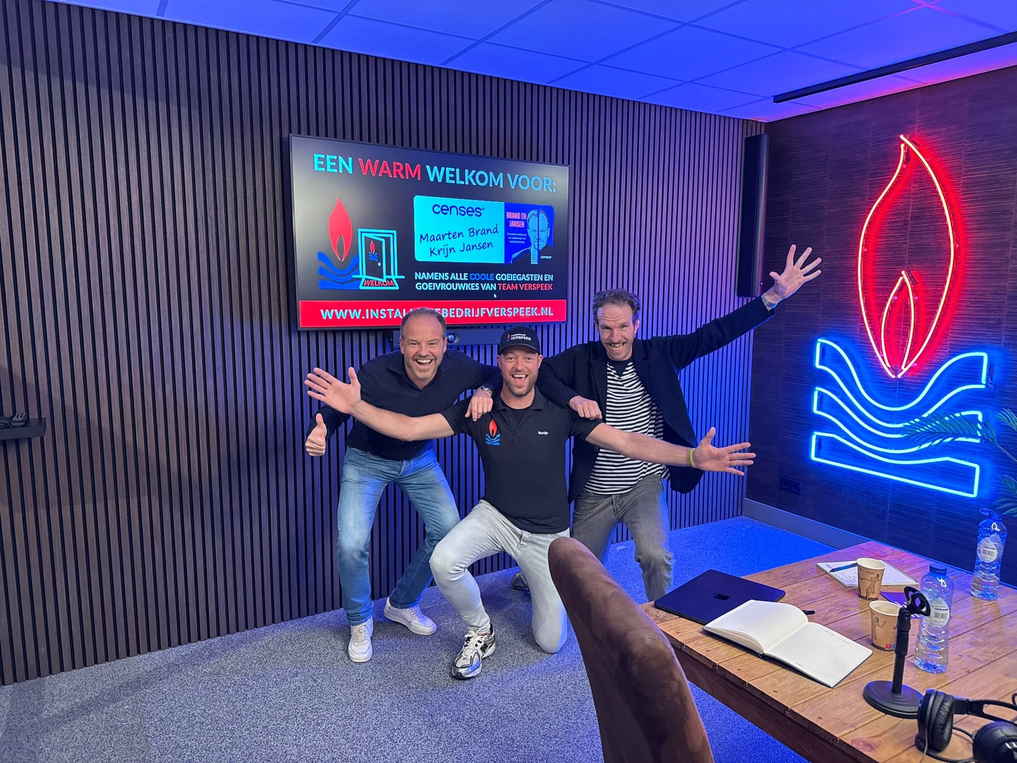 Vijf mensen die blij en enthousiast poseeren in een vergaderruimte, met neonlichten en een scherm op de achtergrond. Bij podcast opname Martijen Verspeek voor Brand en Jansen.