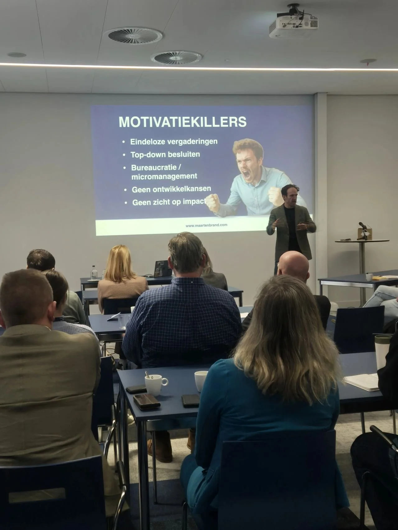 Een presentatie in een conferentiezaal met een projector die een dia toont over Motivatiekillers. De dia bevat een afbeelding van een man die boos is en schreeuwt.