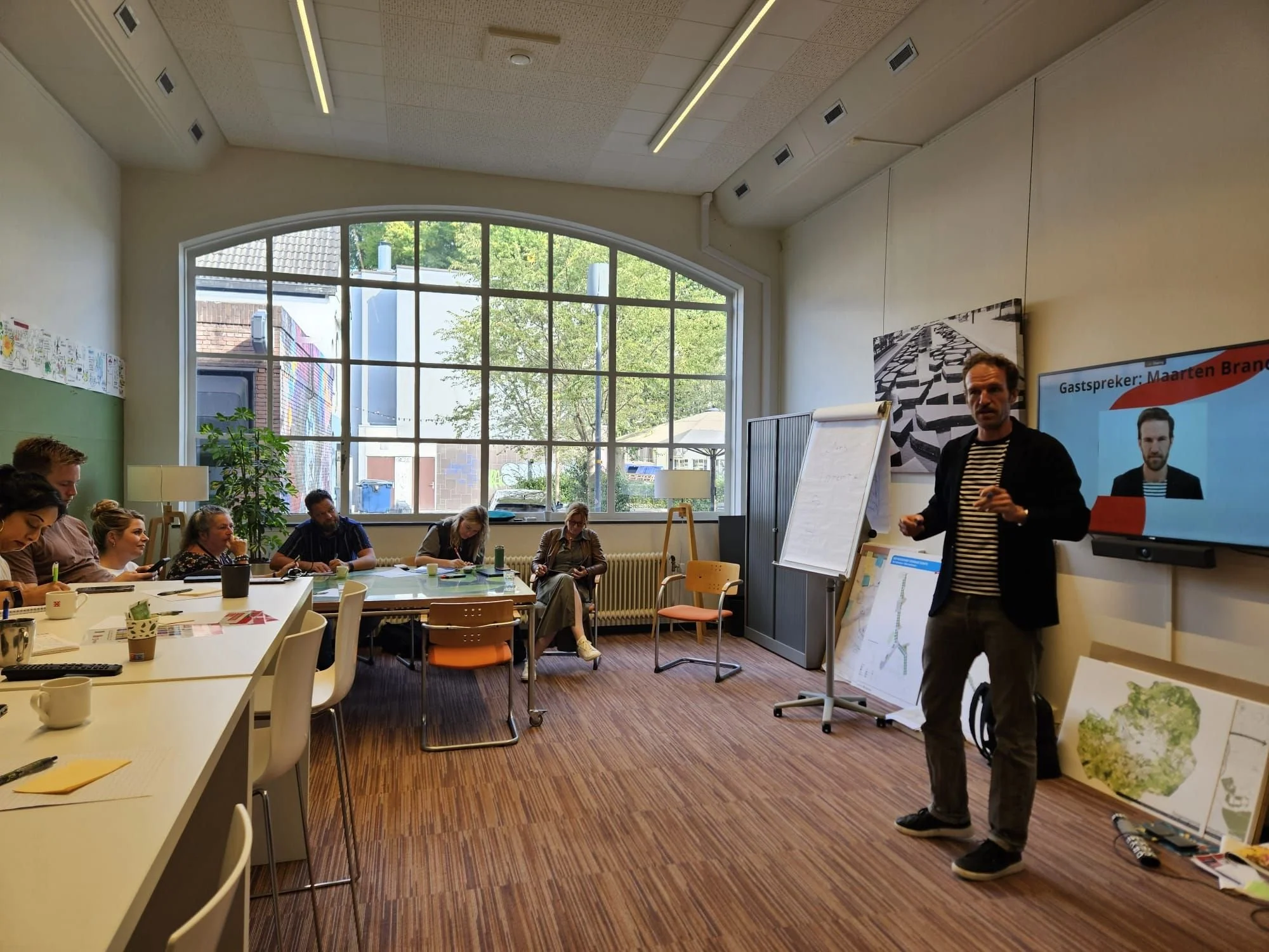 Man geeft presentatie aan een groep mensen in een vergaderruimte met grote ramen, schilderijen en een groot scherm waarop tekst en een afbeelding te zien zijn. Maarten Brand stage-expert, spreker en auteur over praktijkleren