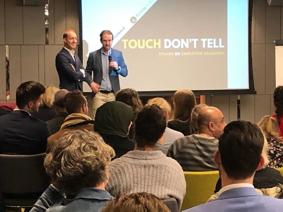 Twee sprekers staan voor een publiek in een conferentiezaal, met een presentatie op de achtergrond die zegt 'TOUCH DON'T TELL' en 'STAGES EN EMPLOYER BRANDING'.