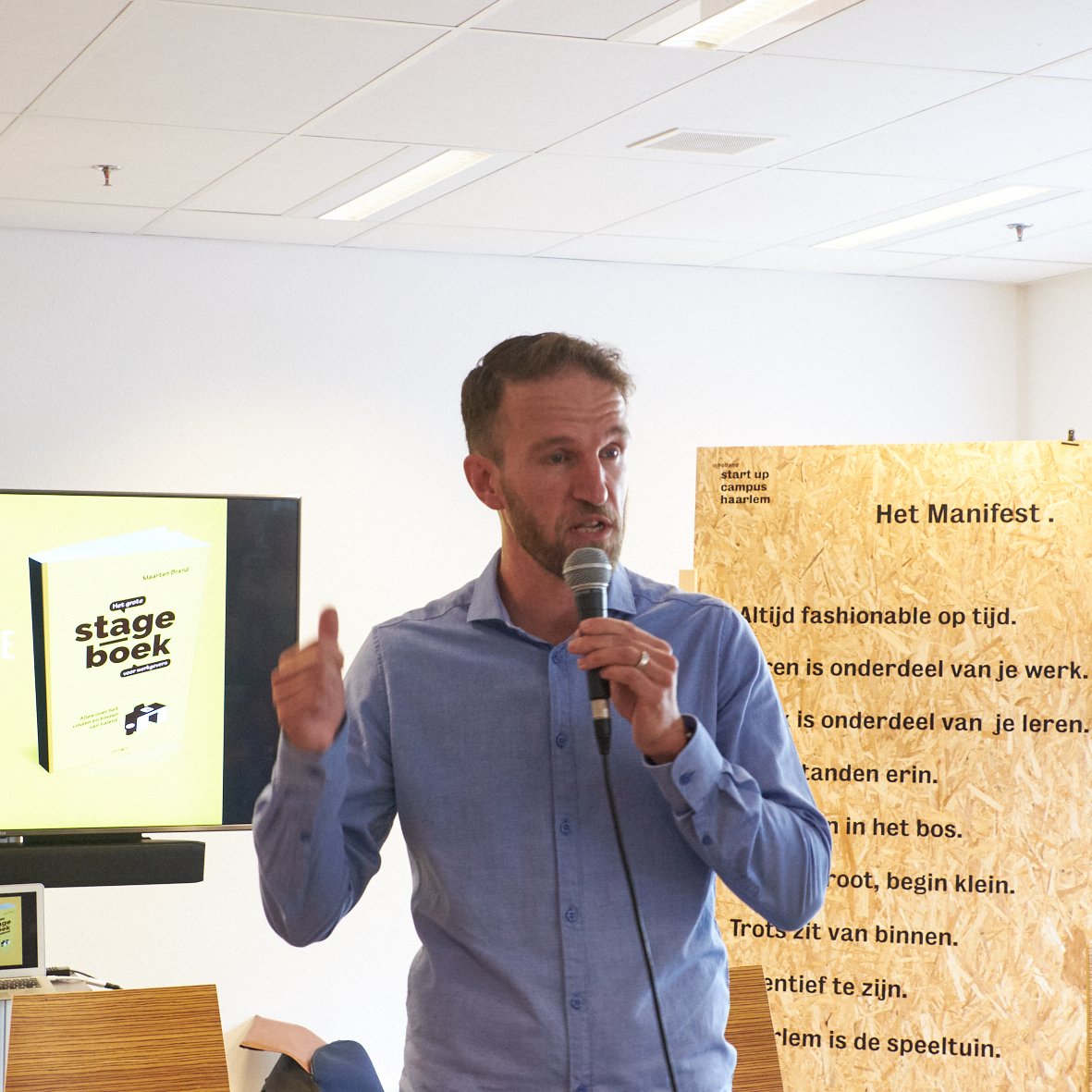 Man houdt een microfoon vast tijdens een presentatie, er zijn posters en een scherm op de achtergrond. Maarten Brand stage-expert, spreker en auteur over praktijkleren