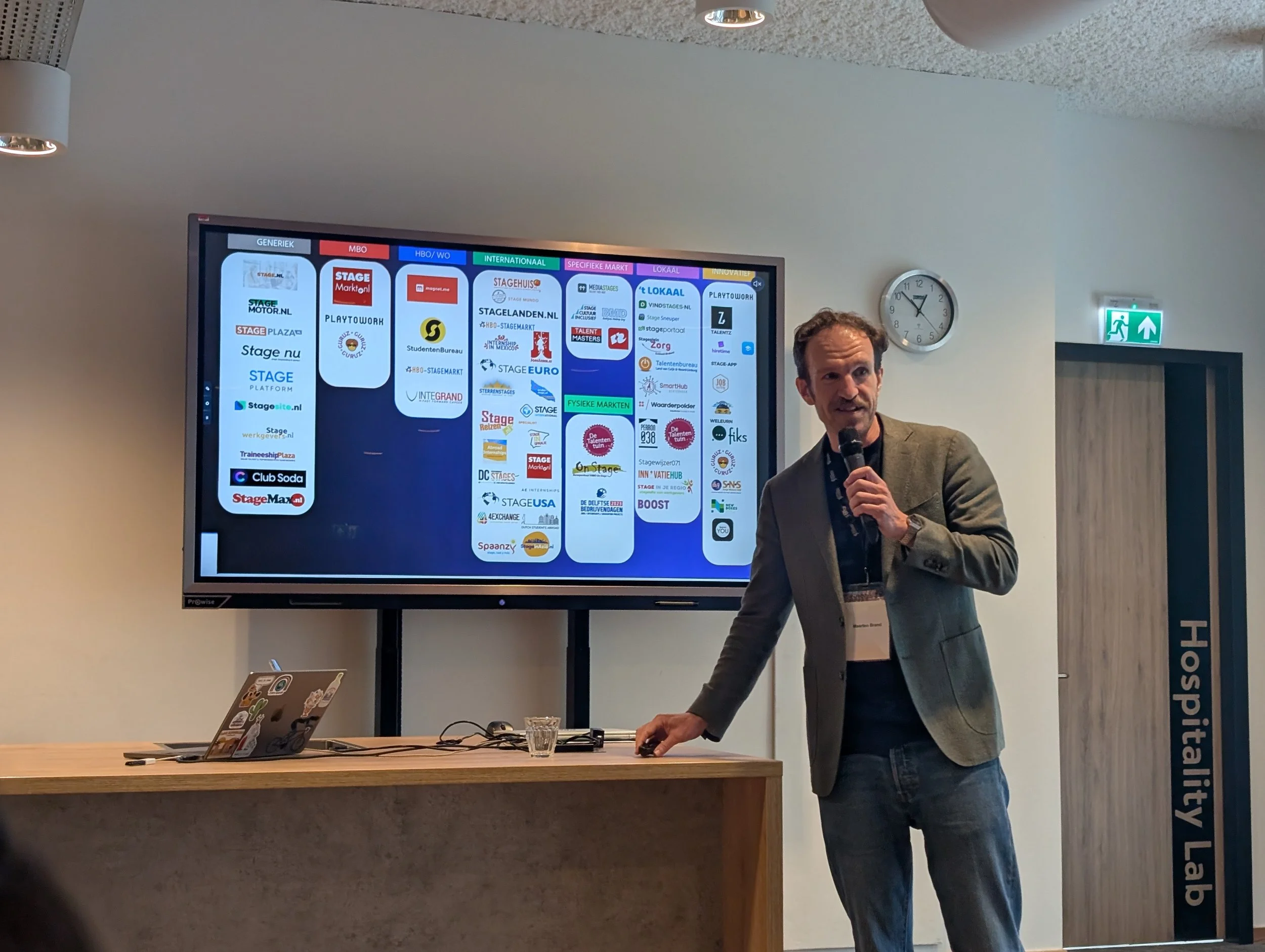 Man die een presentatie geeft voor een scherm met verschillende logo's en tekst, in een zaal met een klok en een uitzichtdeur.