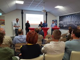 Een groep mensen zit in een zaal en kijkt naar een presentatie met vier verslaggevers of sprekers aan de voorkant.