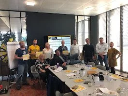 Een groep mensen staat in een vergaderruimte, rondom een lange tafel met ontbijtgerei en documenten, tijdens een bijeenkomst of presentatie. Maarten Brand geeft training 