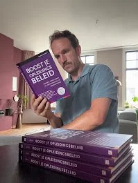 Man die een boek vasthoudt en meerdere kopieën van hetzelfde boek op tafel heeft liggen. Het boek heeft een paarse omslag.