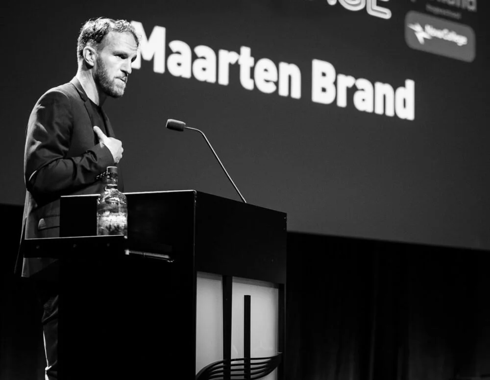 Man staat bij een lessenaar met een microfoon en een fles water, op een podium tijdens een presentatie. Op de achtergrond staat een groot scherm met de tekst 'Maarten Brand'.
