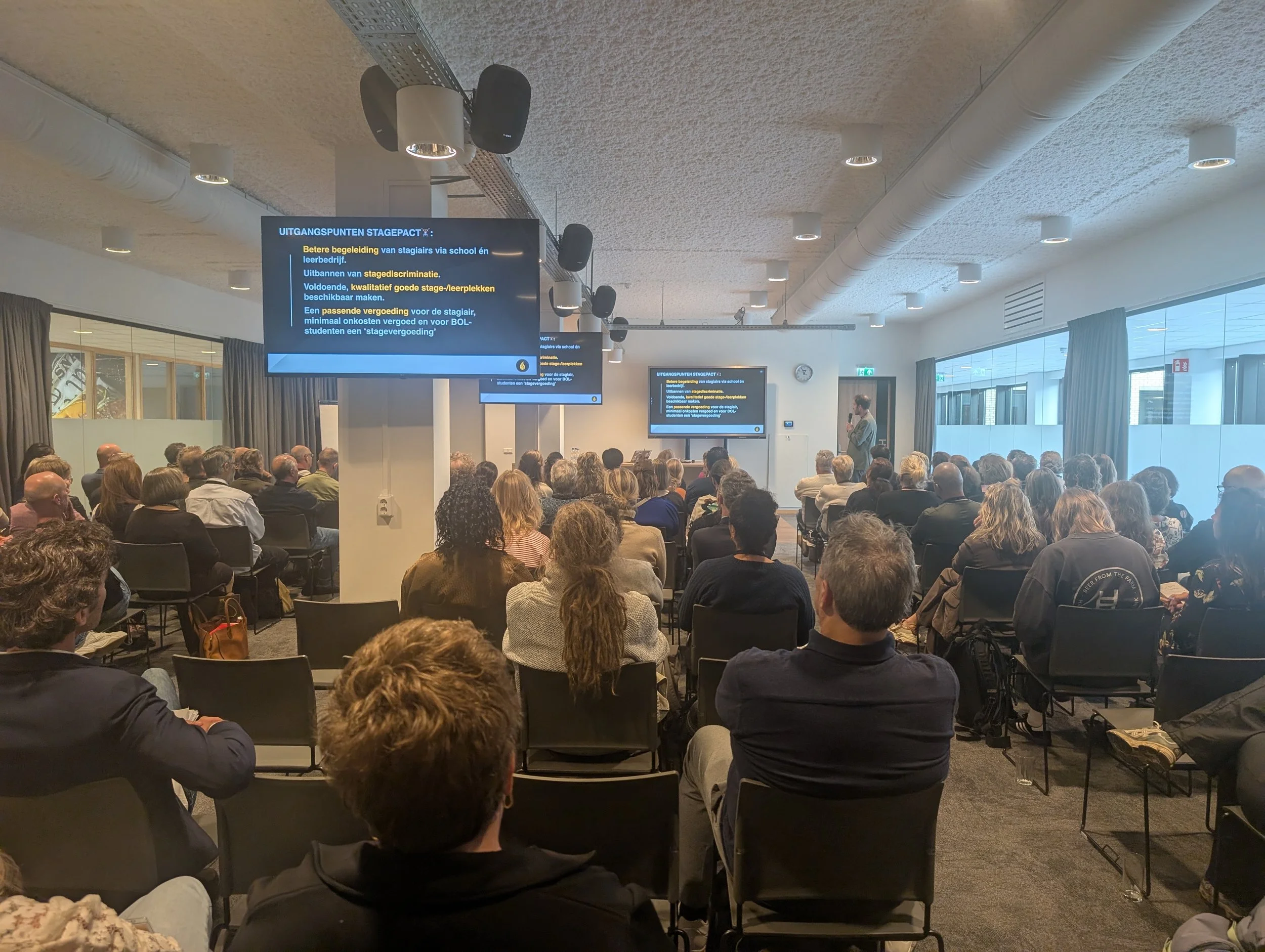Een grote groep mensen zit in een zaal en luistert naar een spreker die voor een scherm staat. Op de schermen wordt een presentatie getoond met tekst in het Nederlands.