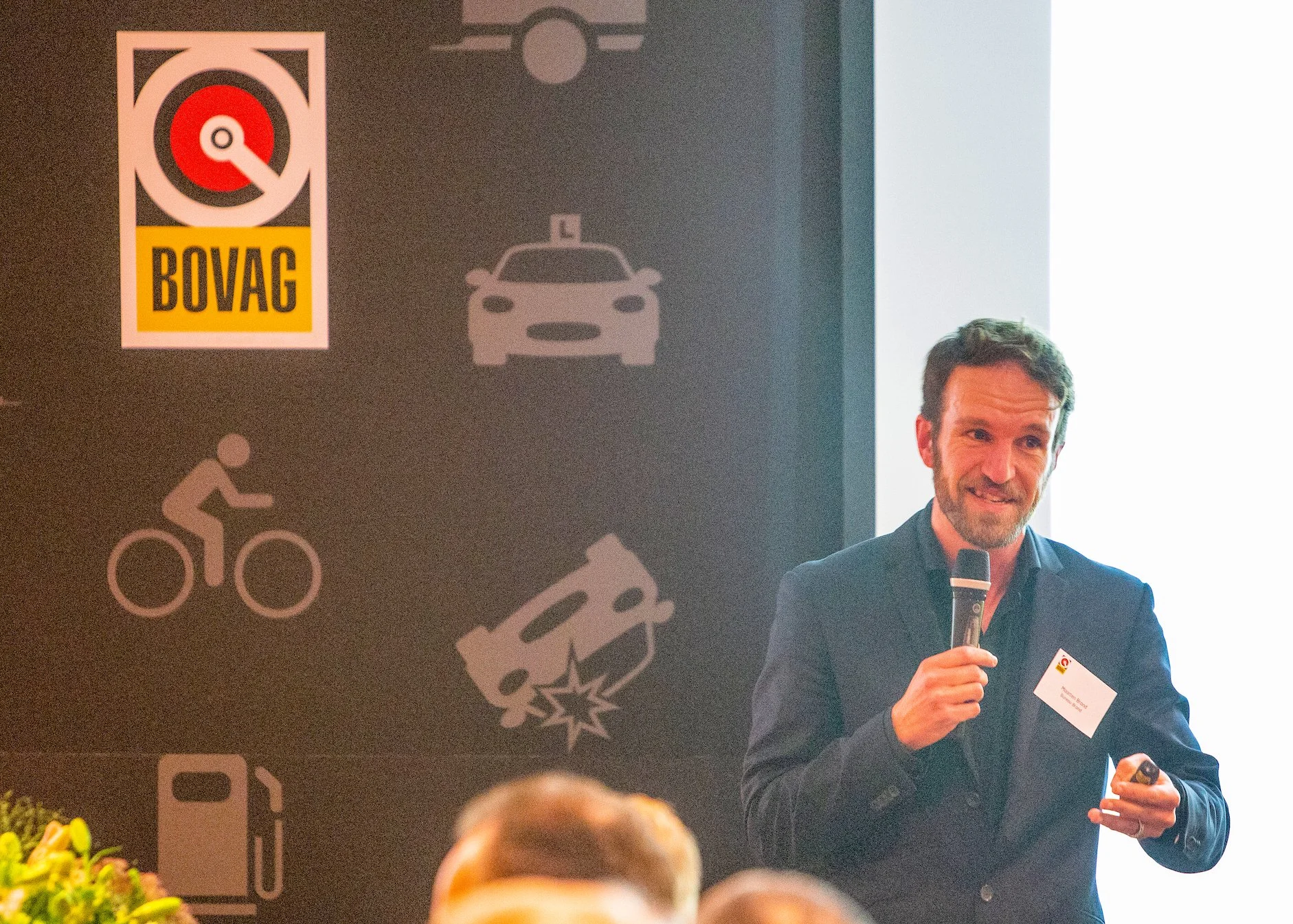 Man die op een podium spreekt met een microfoon, achtergrond met verkeersveiligheidssymbolen en het BOVAG-logo.