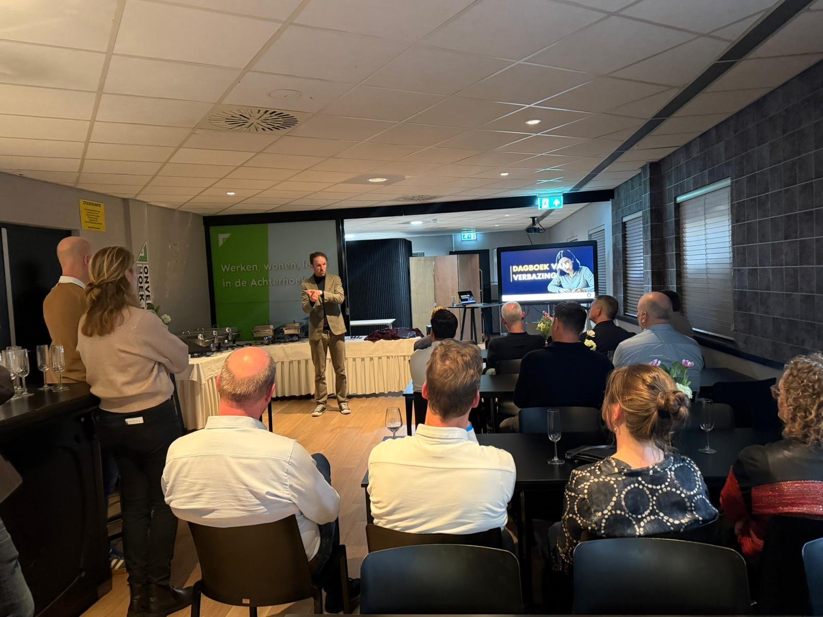 Een man staat voor een groep mensen die naar hem luisteren tijdens een presentatie in een vergaderzaal. Op het scherm op de achtergrond staat 'DAGBOEK VAN VERBAZING' en er bevindt zich een buffet aan de zijkant Maarten brand stage-expert..