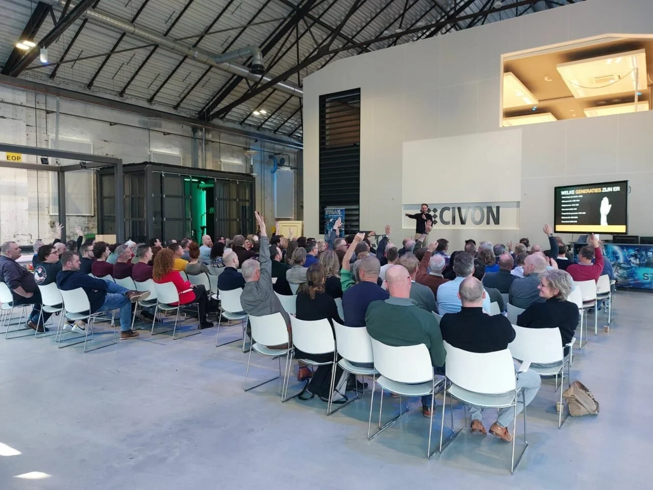 Een grote groep mensen zit in een conferentiehal, luisterend naar een spreker die staat voor een scherm en een muur met het logo 'CIVON'. Meerdere mensen steken hun hand op tijdens de presentatie. Maarten Brand stage-expert, spreker en auteur over pr