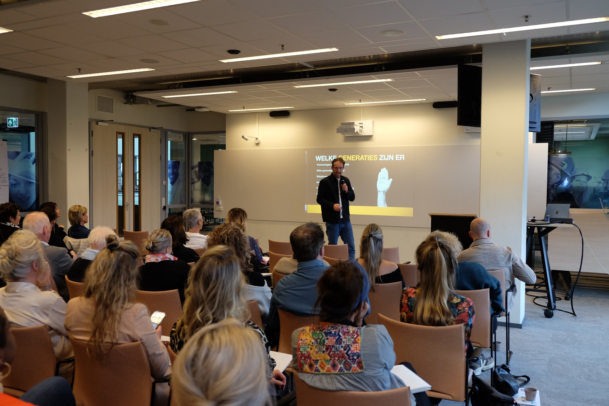 Een man geeft een presentatie voor een zaal met luisteraars. Op de achtergrond staat een scherm met tekst en een afbeelding van een hand. De zaal is gevuld met mensen die naar de spreker kijken. Maarten Brand stage-expert, spreker en auteur stages