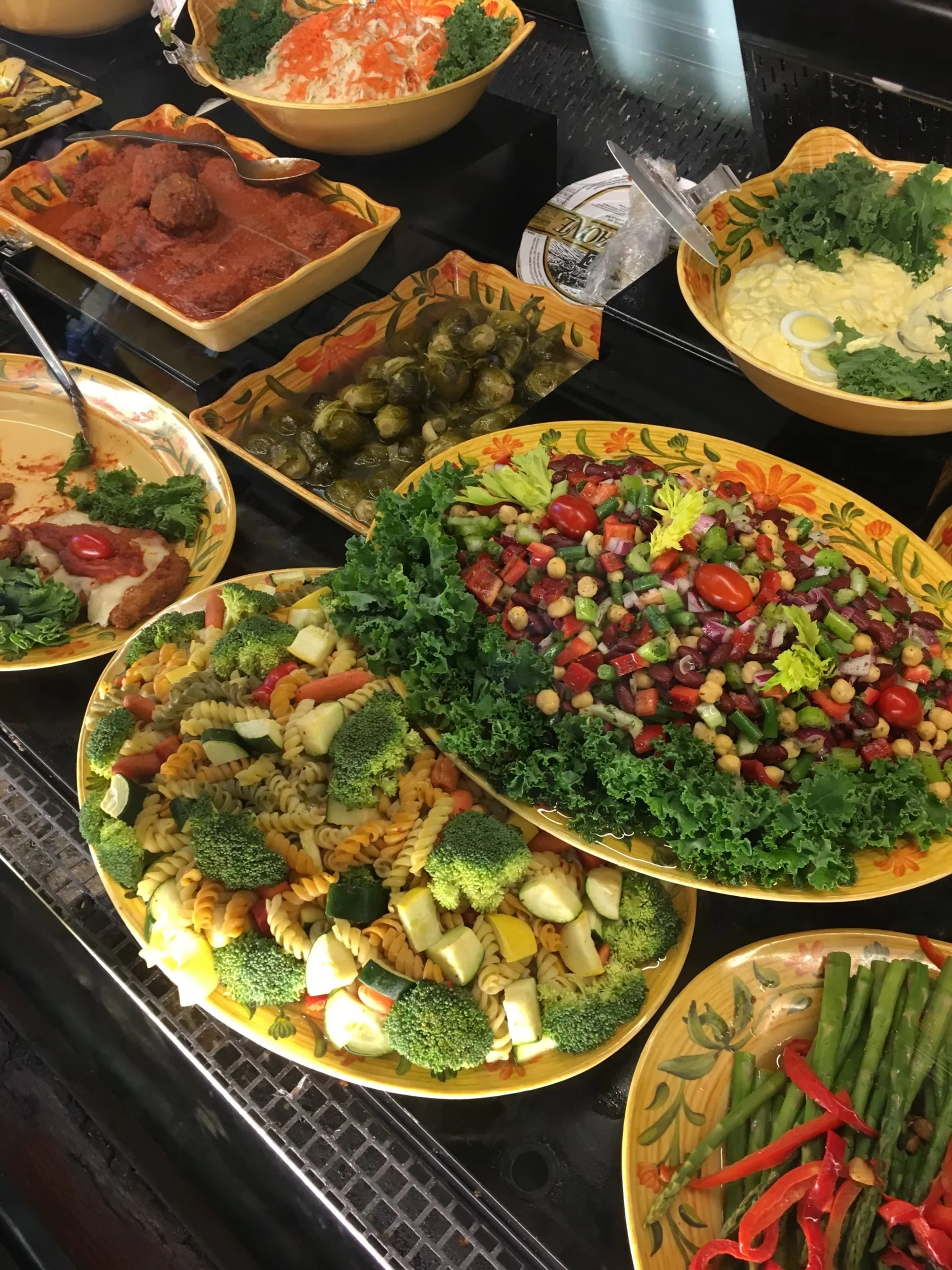 A buffet table with various colorful salads and dishes, including pasta salad with broccoli and zucchini, a vegetable salad with cherry tomatoes, green beans with red peppers, and other assorted vegetables, all served on decorative yellow plates.