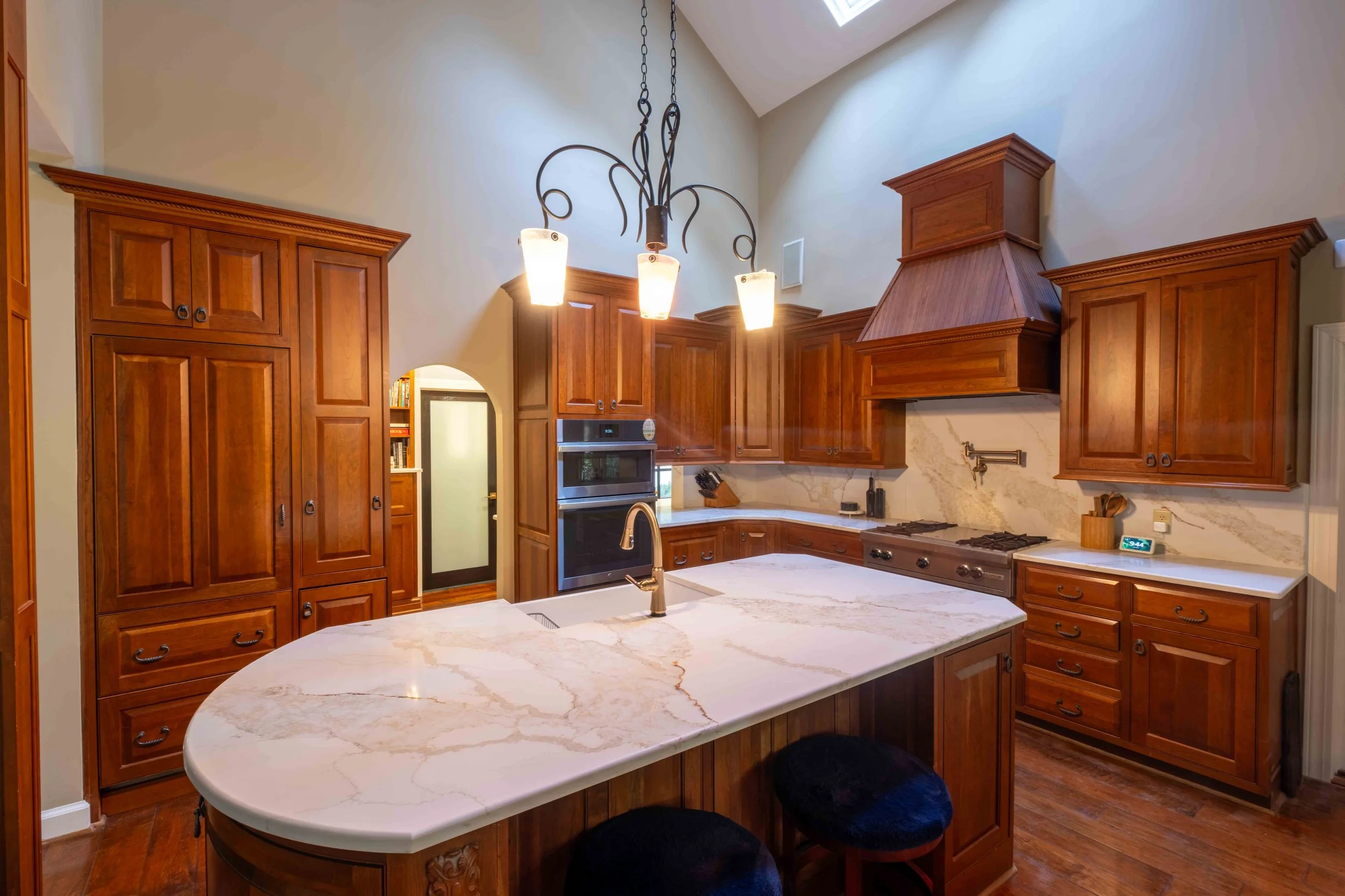 Kitchen with wooden cabinets, marble countertop island, stainless steel oven, range hood, and a hanging light fixture.