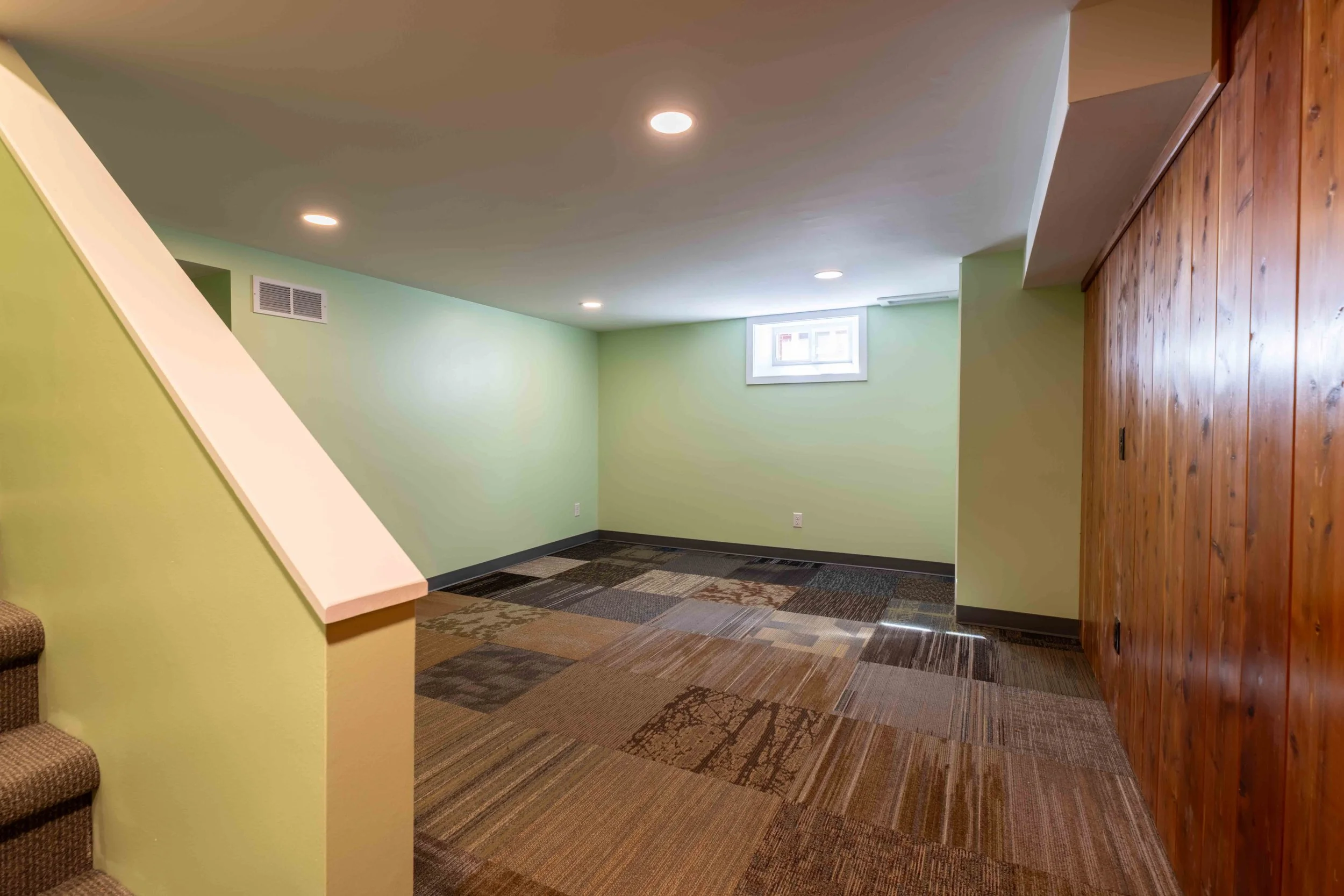 Interior view of a room with light green walls, a small window, a wooden sliding door, carpeted floor, and recessed ceiling lights.
