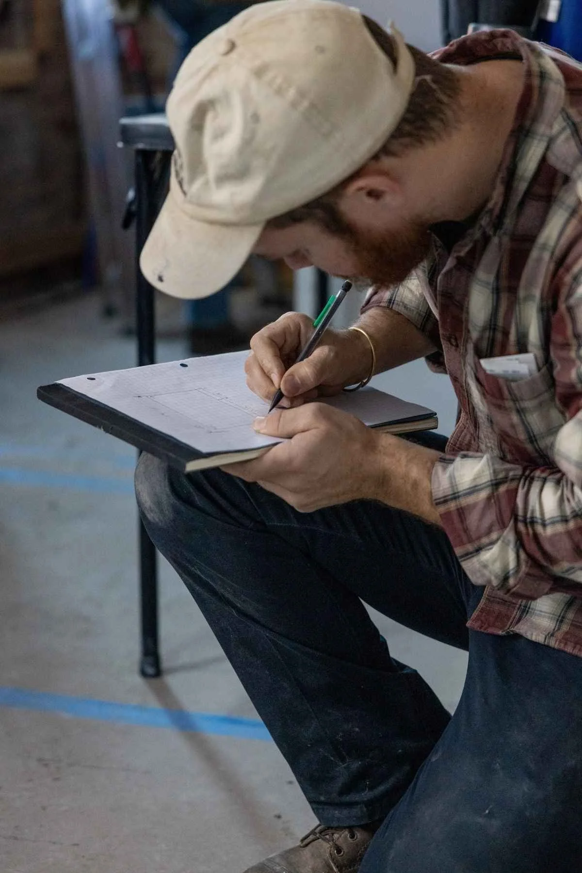 A man wearing a baseball cap and plaid shirt is sitting on a chair, writing on a piece of graph paper with a green pen. The background appears to be a workshop or storage room.