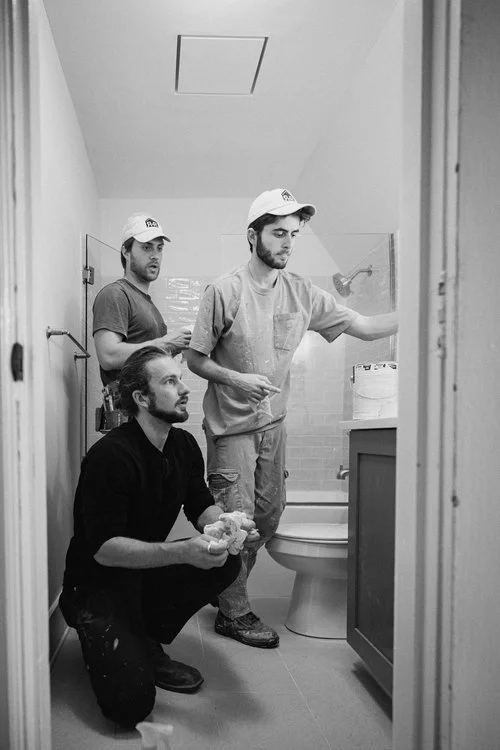 Three men working in a bathroom, one is kneeling with a rag, another is standing near a toilet, and the third is in the background near the shower.