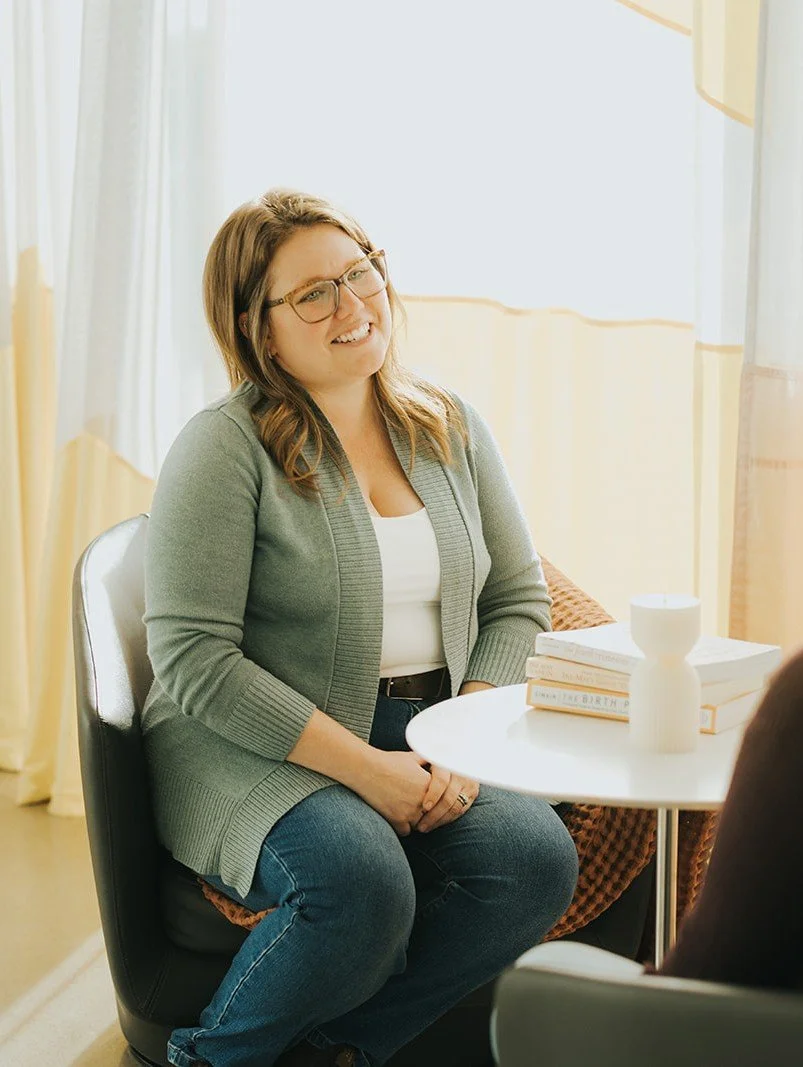 A woman with glasses, wearing a gray cardigan and jeans, sitting at a table in a bright room, engaging in a conversation.