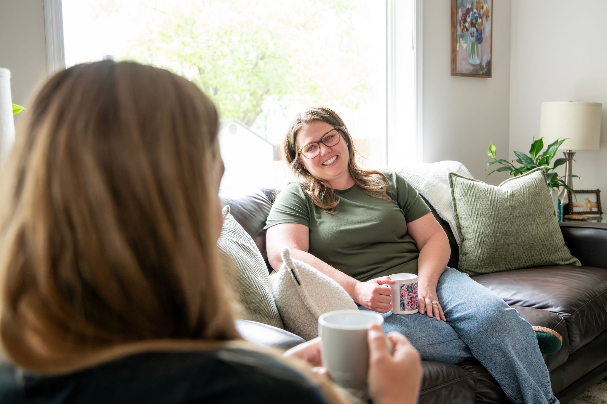 Two women sitting on a couch having a conversation, one is smiling and holding a mug, in a well-lit living room with green plants and artwork on the wall.