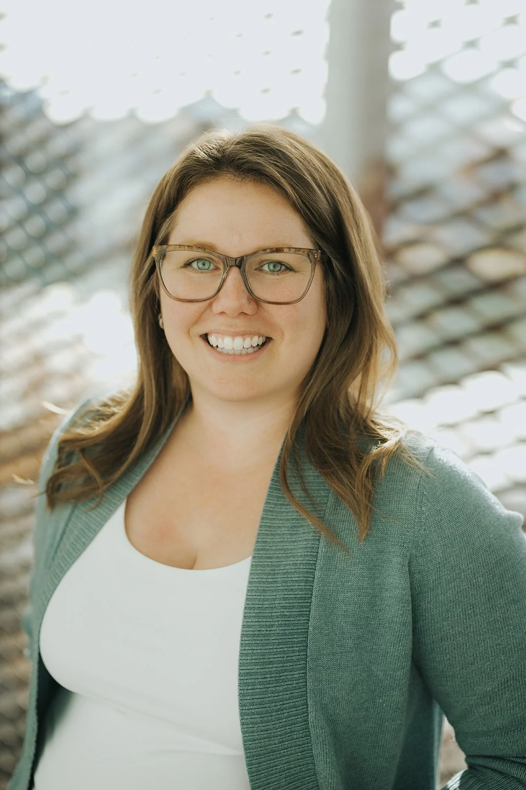 A woman with light skin, brown hair, and glasses smiling outdoors, with a wooden railing in the background.
