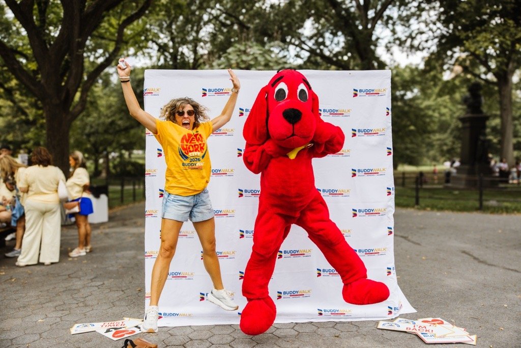 a woman wearing a yellow shirt, shorts and sunglasses jumps with a huge smile beside a large costumed person jumping while dressed as Clifford the Big Red Dog. They are in front of a Buddy Walk step and repeat background with a canopy of trees.