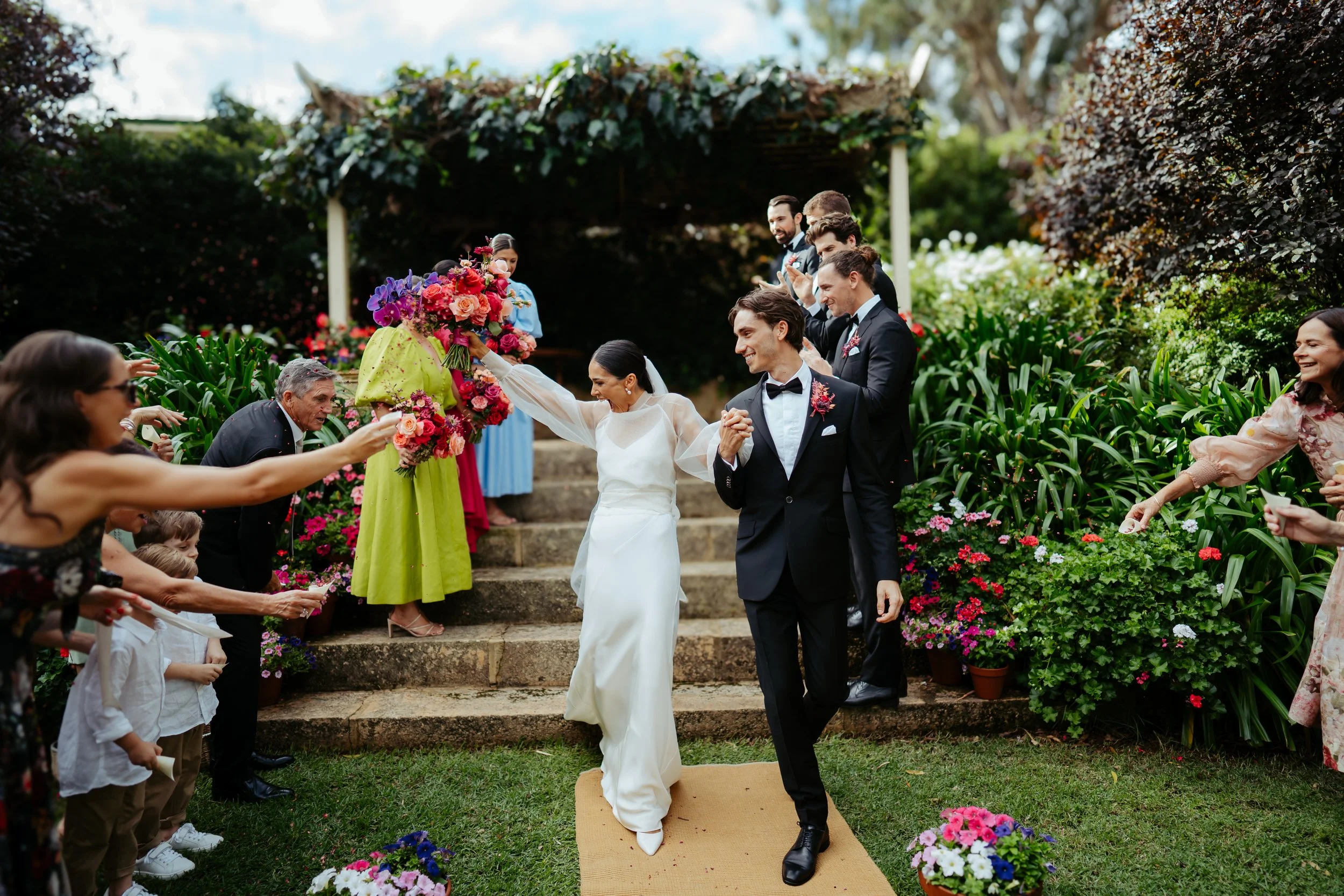 A newly married couple walking down the aisle outdoors, surrounded by friends and family throwing flowers, with a lush garden backdrop and cloudy sky.