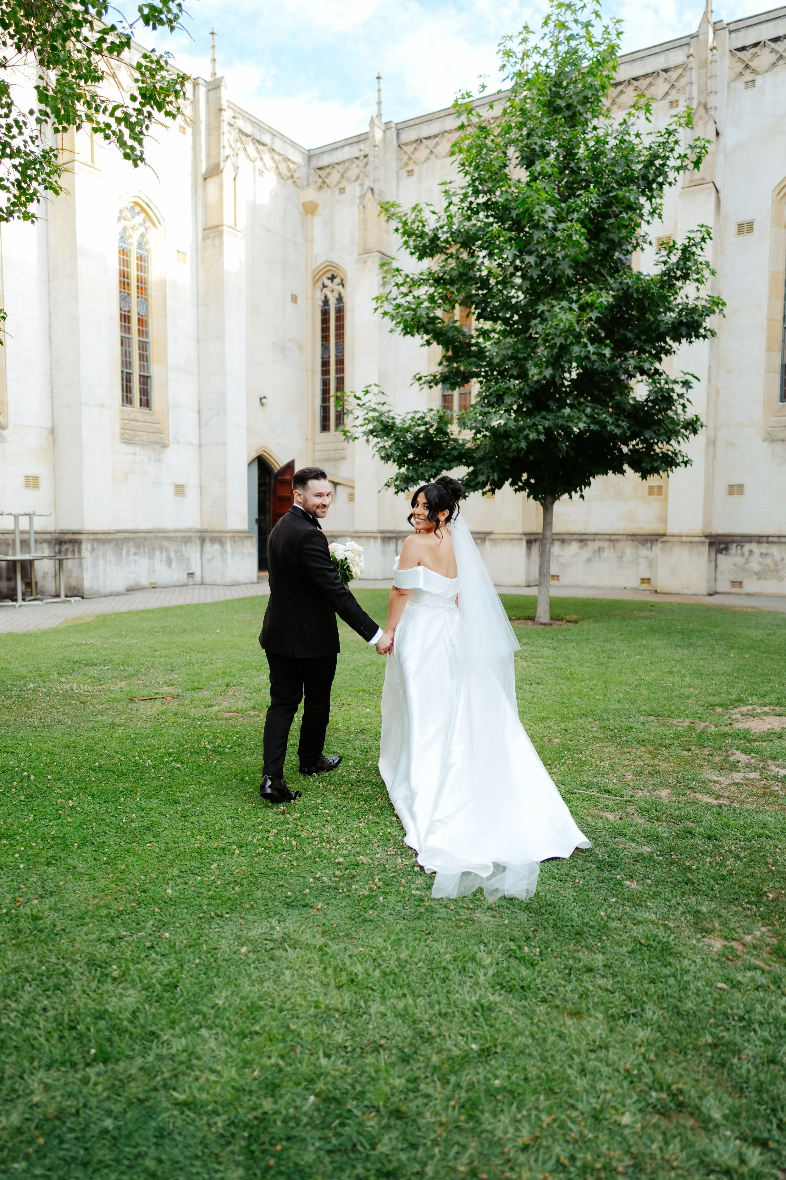 A bride and groom holding hands and smiling at each other in front of a large tree and a historic church building with stained glass windows.
