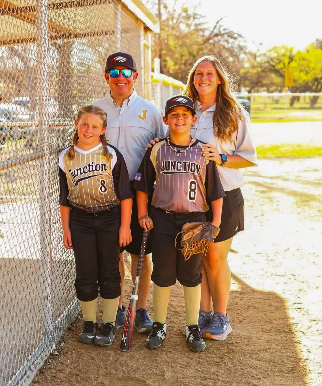 A family of four standing at a baseball field, dressed in baseball uniforms, smiling for a photo.