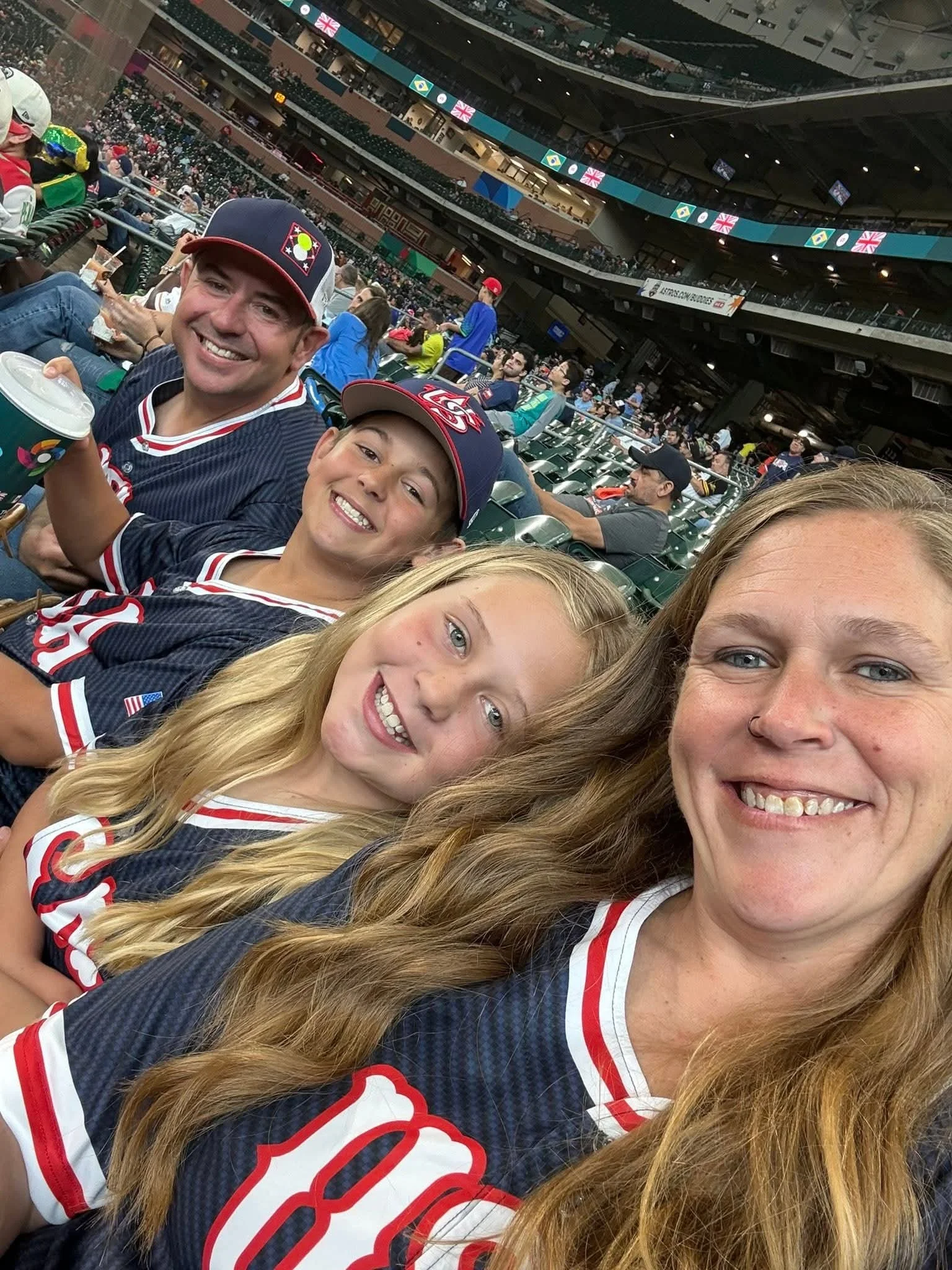 Group of baseball fans sitting in a stadium, wearing team jerseys and caps, with some holding drinks, while watching a game.