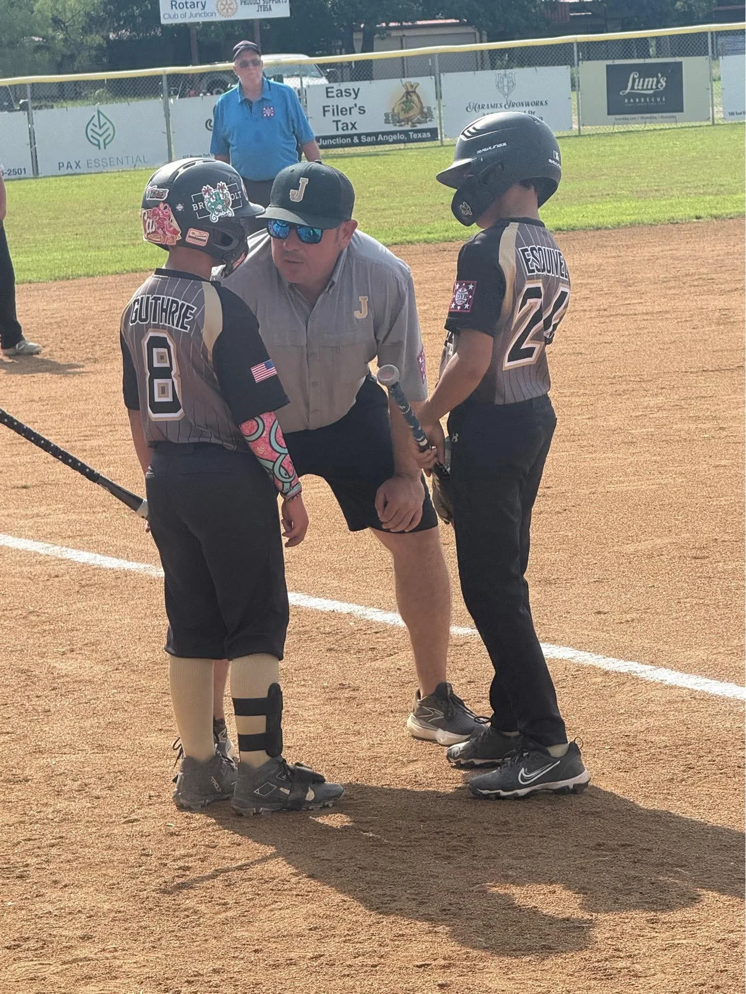 A person holding a baseball bat, surrounded by two children dressed in baseball uniforms and helmets, with a coach or referee talking to them on a baseball field.