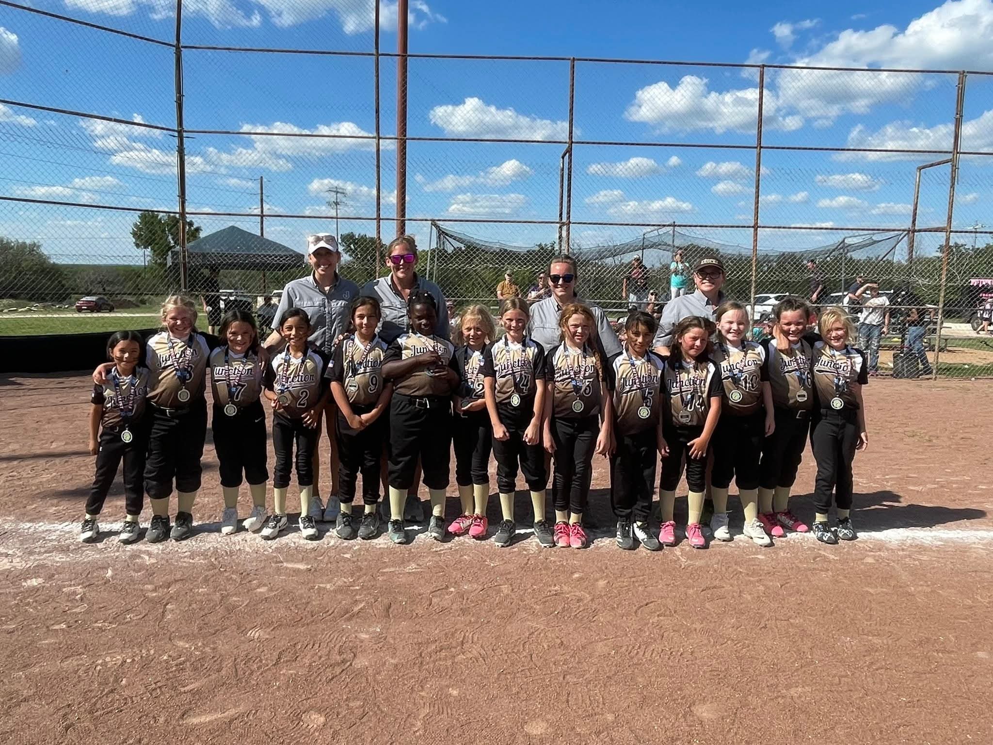 A group of young girls and their coach posing on a baseball field during daytime. The girls are wearing matching jerseys and medals. The background features a chain-link fence, a tent, cars, and a partly cloudy sky.