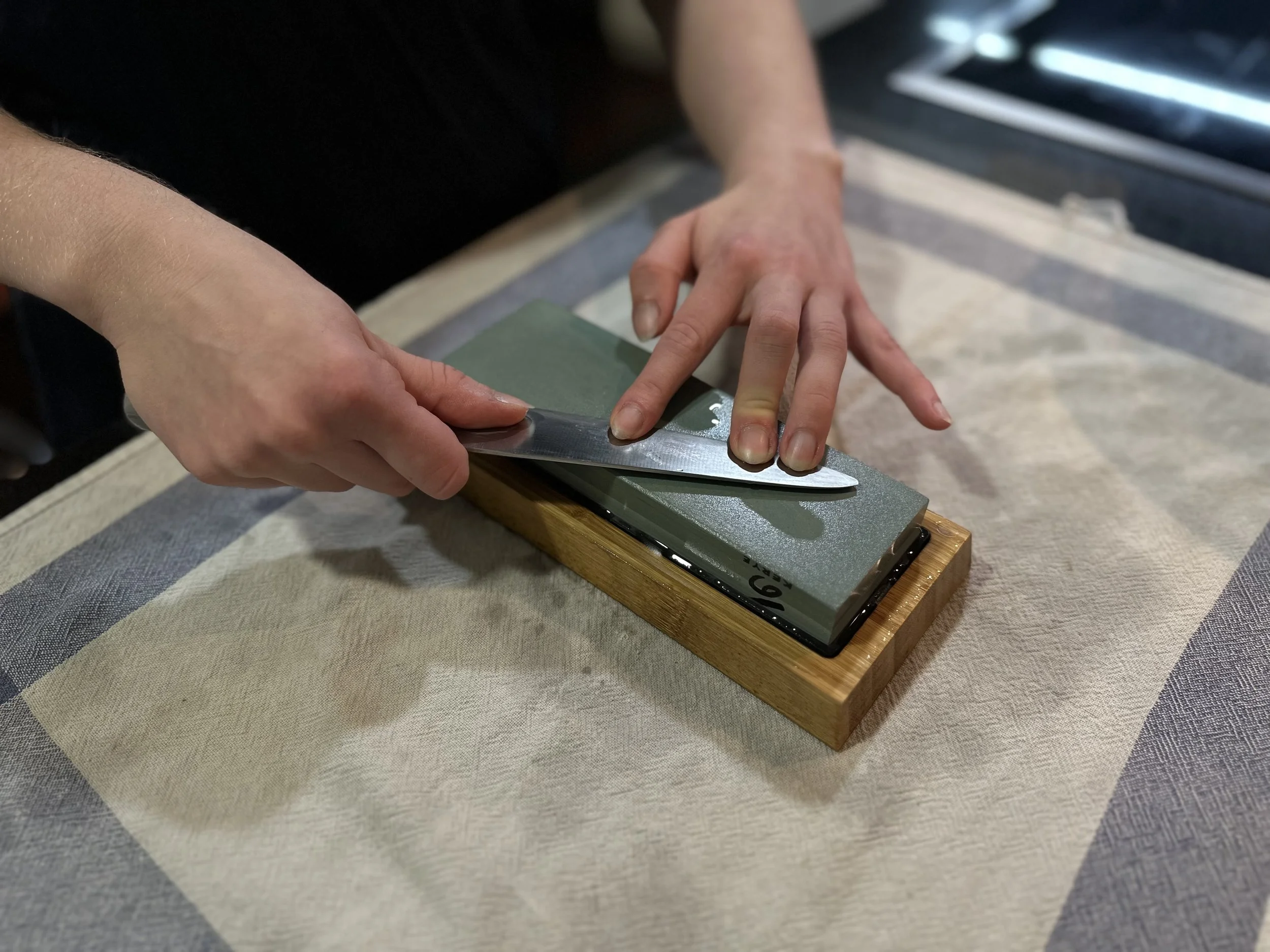 A white and blue towel on a black countertop with a green sharpening stone held in place by a wooden block. Two female hands are sharpening a knife on top.