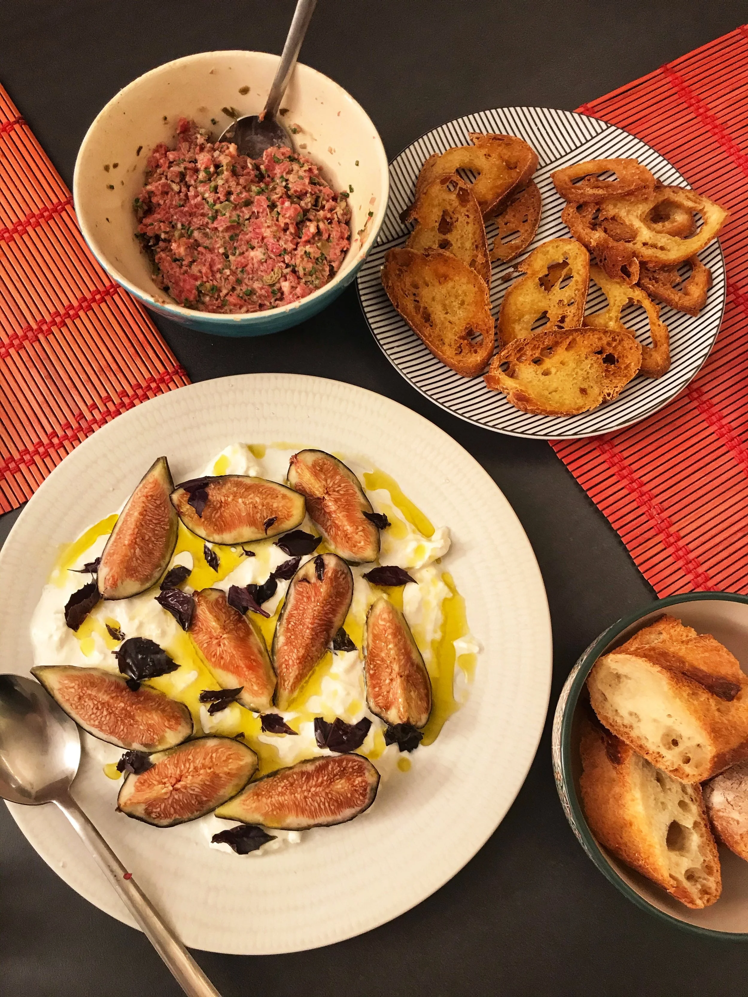 Burrata with figs, steak tar tar, and assorted breads on a grey table with red place mats