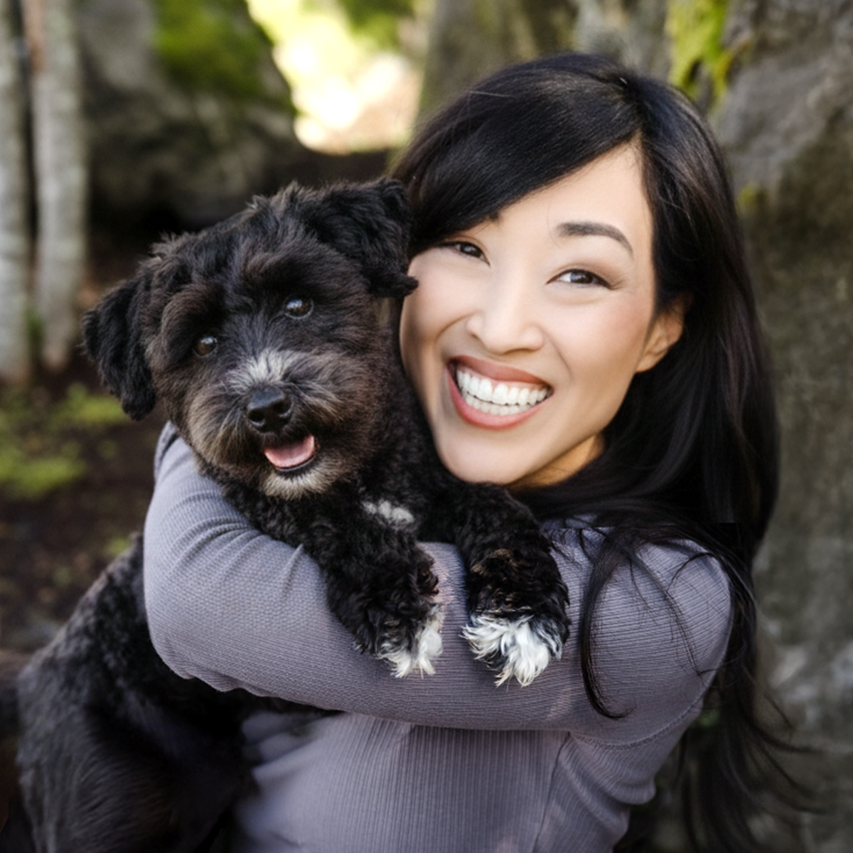 A close-up, outdoor portrait of a young adult woman embracing a small, black Miniature Schnauzer