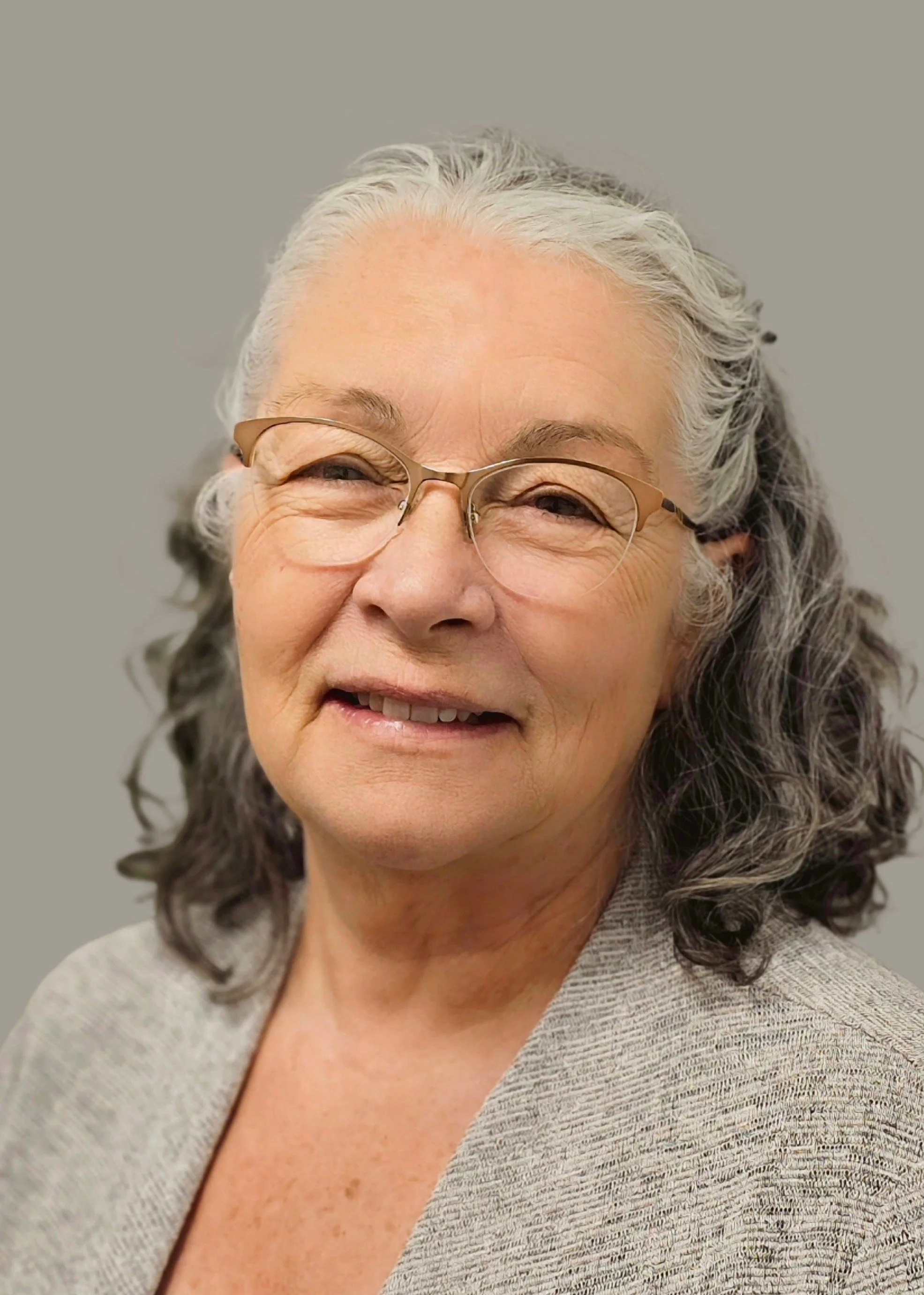 Close-up portrait of an elderly woman with gray hair, wearing glasses and a beige blazer, smiling gently against a plain gray background.
