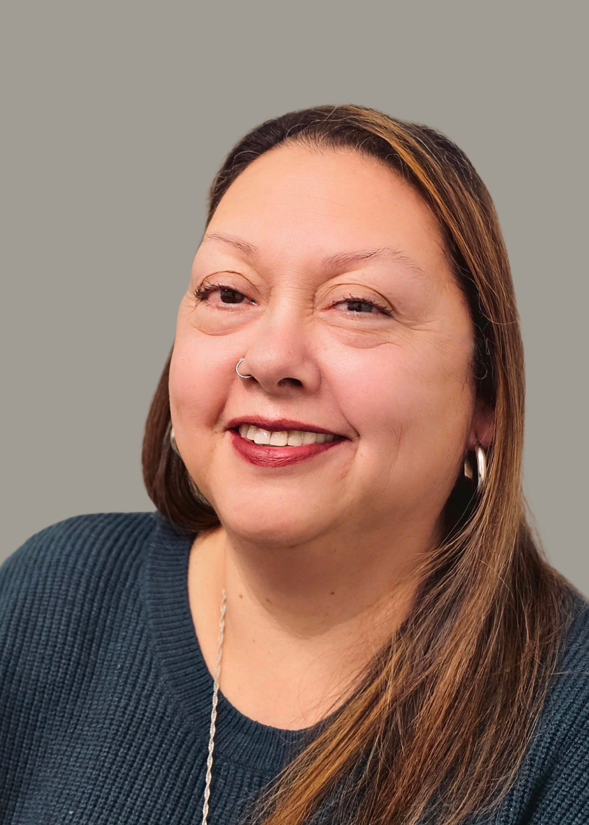 A woman with long brown hair, a nose ring, and hoop earrings, smiling and wearing a dark sweater.