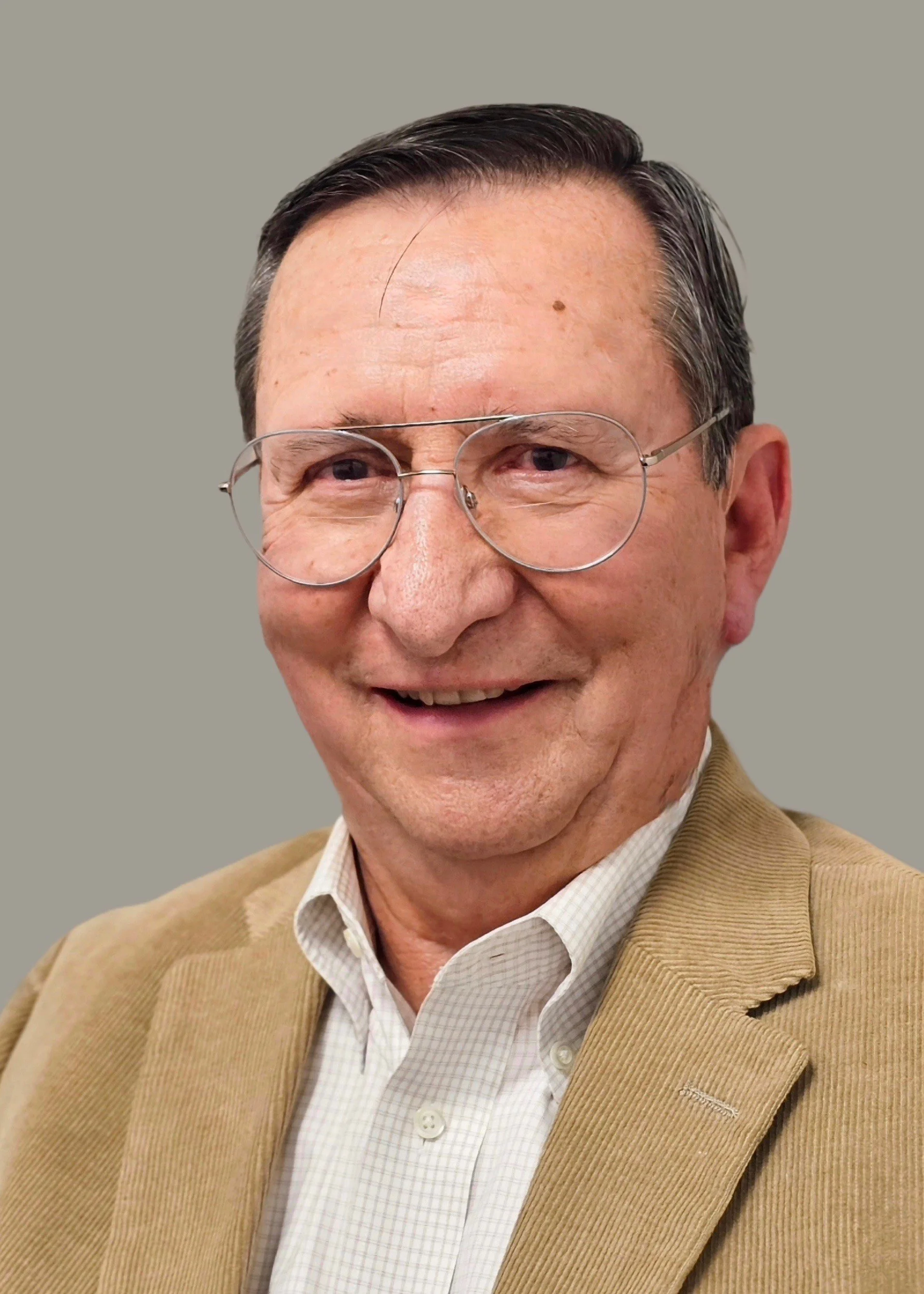 Close-up of an elderly man with glasses, smiling, wearing a beige blazer over a white checkered shirt, against a plain light gray background.