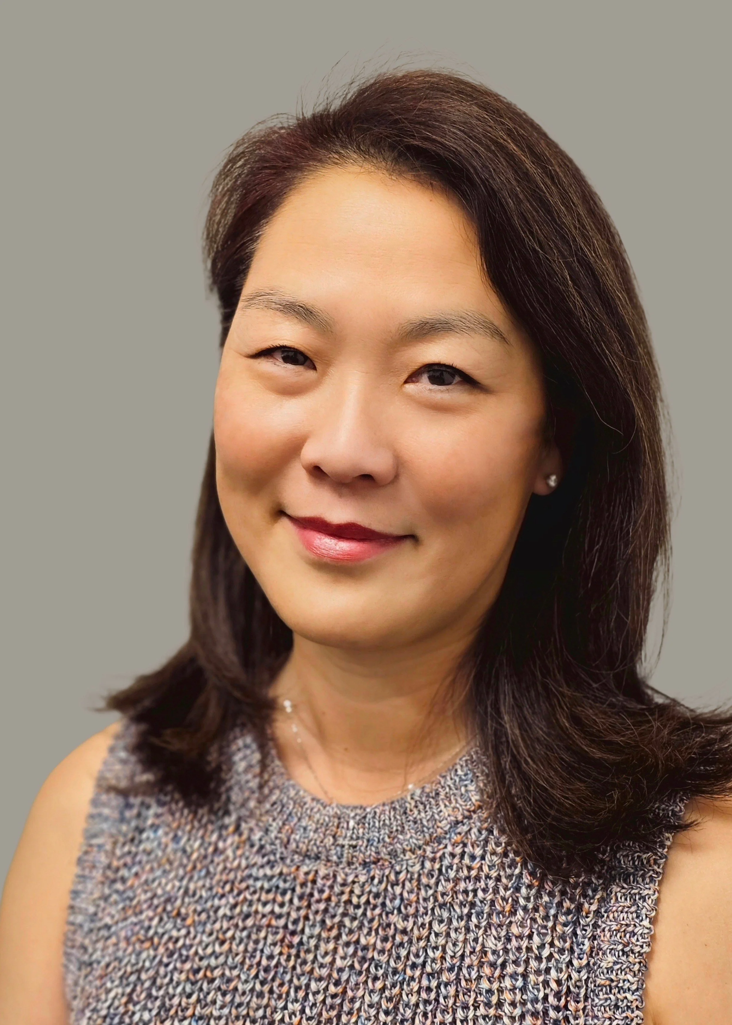 A woman with dark brown hair, fair skin, and a gentle smile, wearing a sleeveless knit top and pearl earrings, posing against a plain gray background.