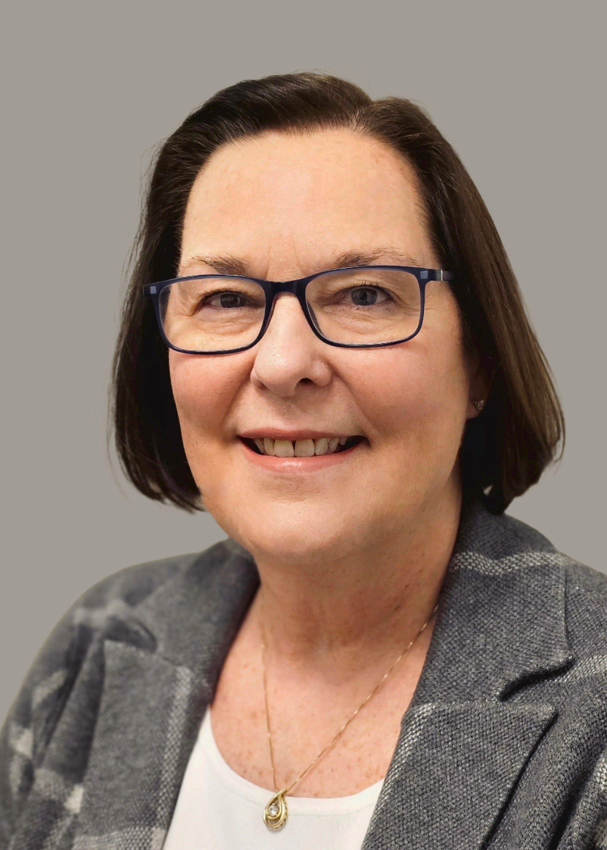 A smiling woman with short dark brown hair, wearing glasses with a dark frame, a gray checkered blazer, a white top, and a gold necklace with a teardrop pendant.