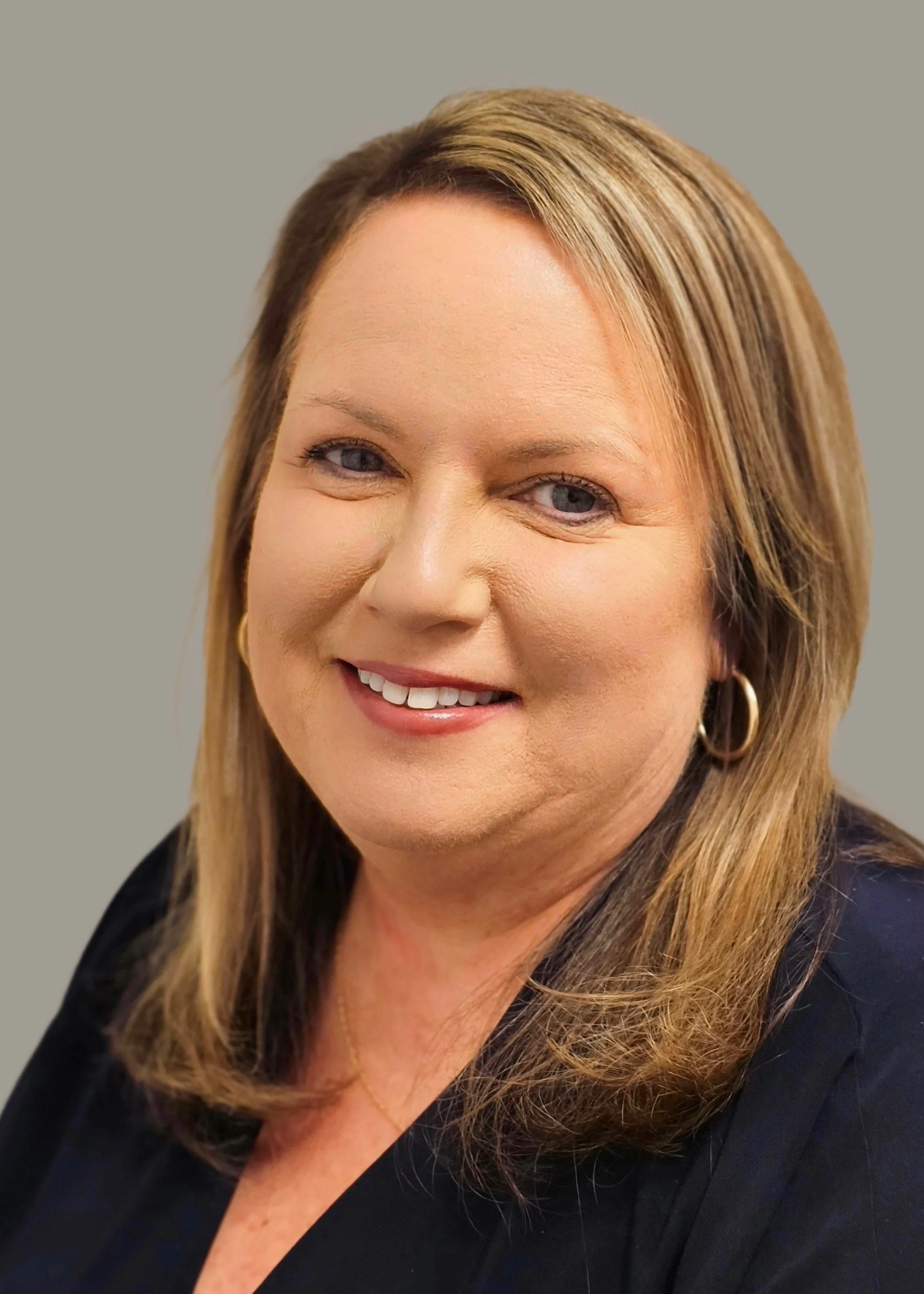 Close-up portrait of a smiling woman with shoulder-length light brown hair, wearing gold hoop earrings and a black top, against a neutral background.