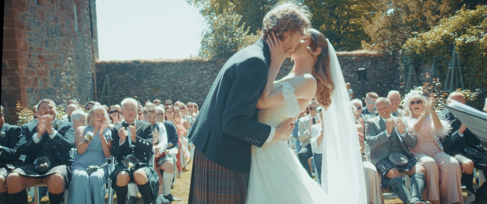 A Bride in a white wedding dress with a long veil and Groom wearing a kilt sharing their first kiss as husband and wife
