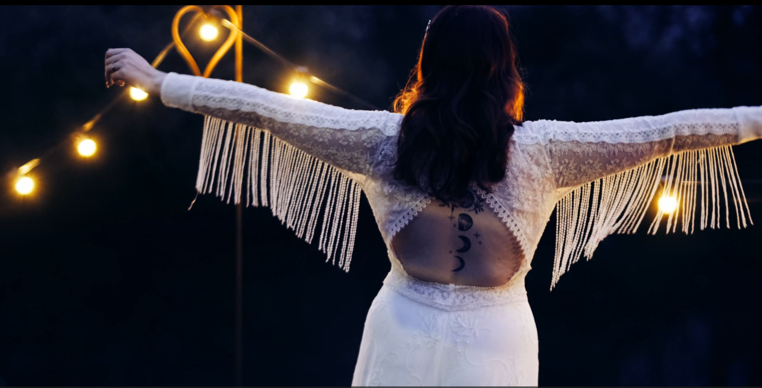 Woman standing outdoors at night with arms outstretched, wearing a white lace dress with fringe sleeves and a tattoo of phases of the moon on her back, illuminated by string lights.