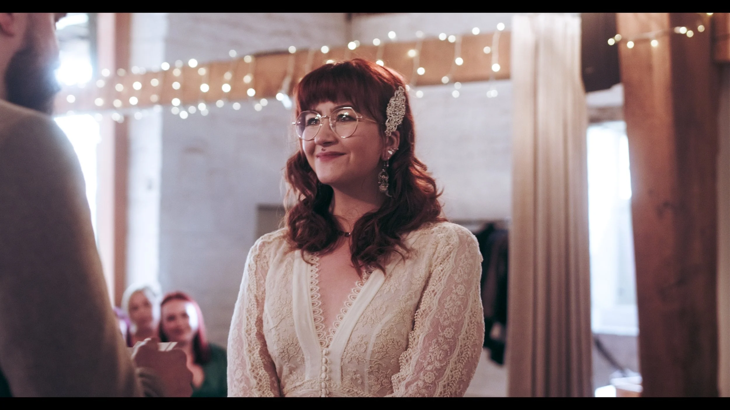 A woman with red hair and glasses, wearing a lace dress, smiling during a wedding ceremony with fairy lights in the background.