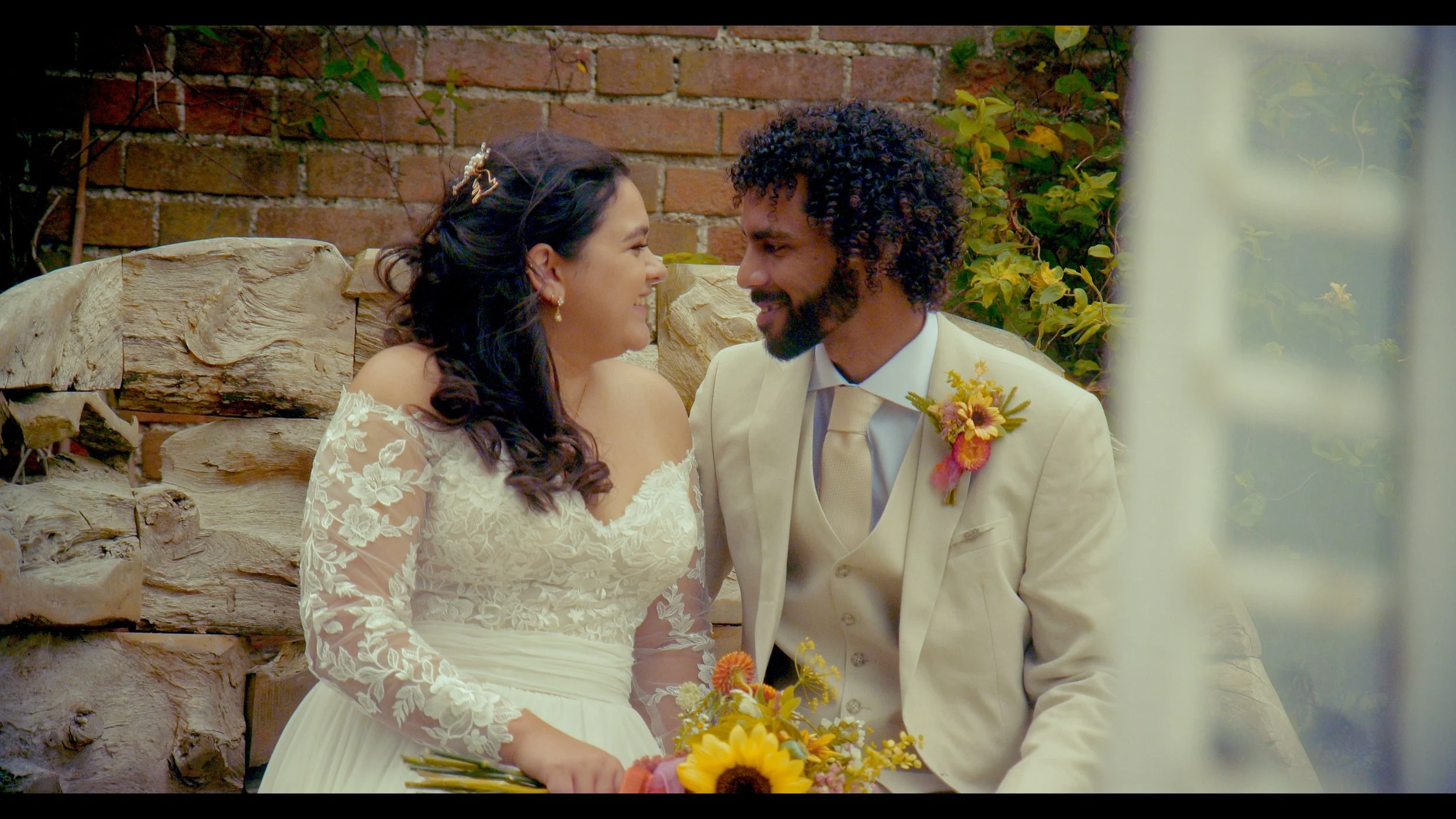 A bride and groom sitting close, smiling at each other during a wedding ceremony outside. The bride is wearing a lace wedding dress with floral embroidery, and the groom is in a cream suit with a boutonniere featuring yellow and pink flowers. The bac