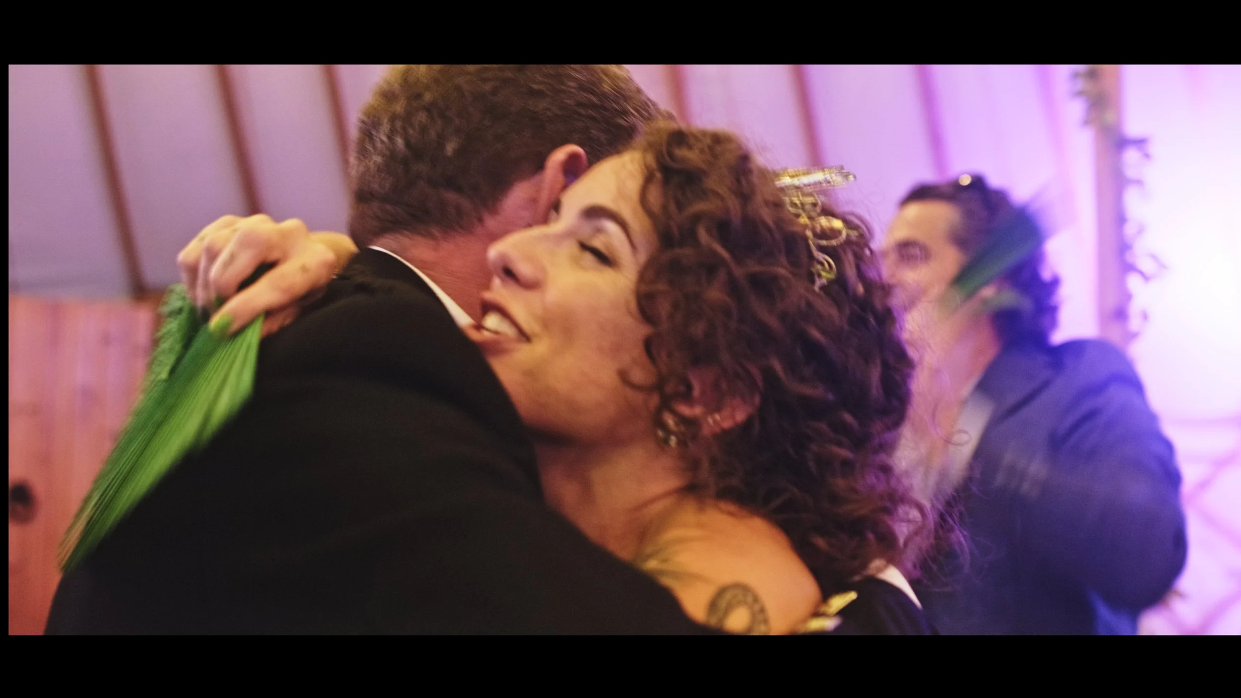 People hugging at a celebration or party, one woman with curly hair and a man with short hair, both smiling.
