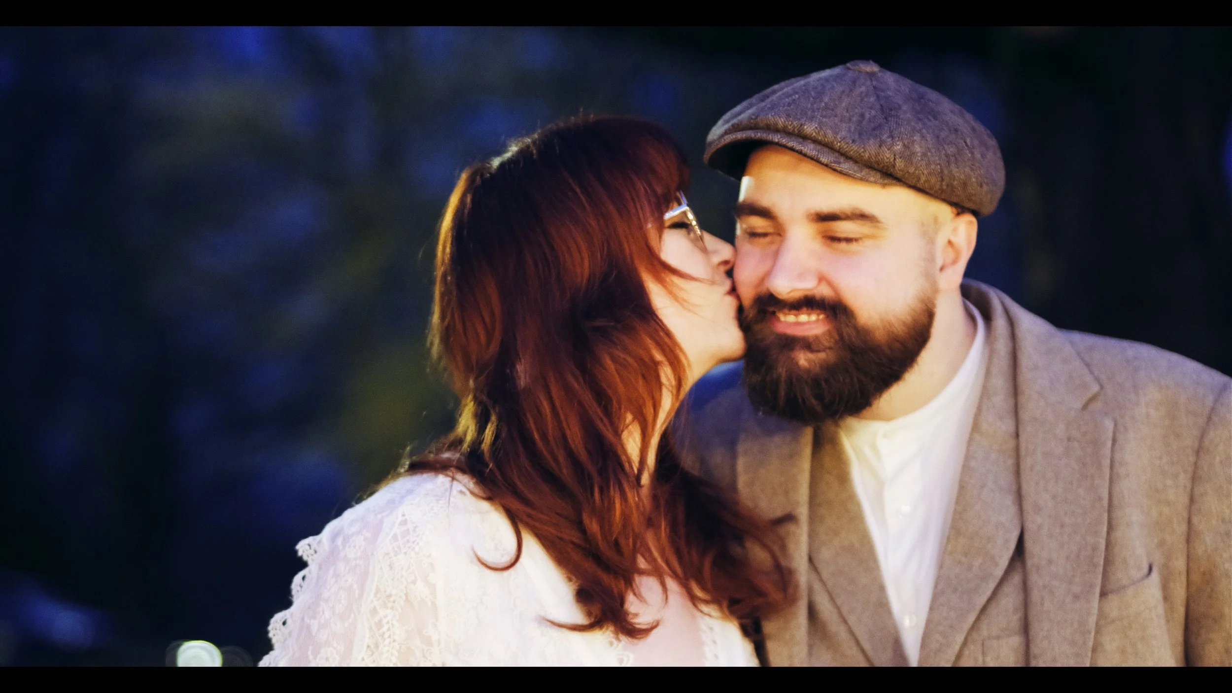 A woman with red hair and glasses kisses a smiling man with a beard, wearing a gray flat cap and a beige blazer, at dusk or evening outdoors.