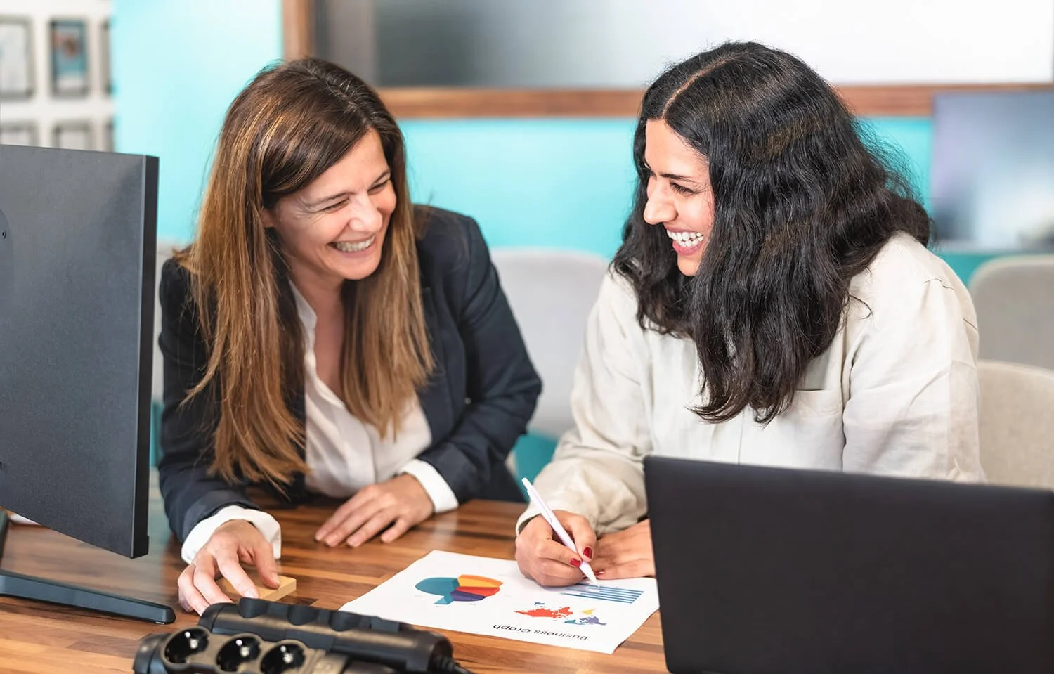 Deux femmes souriantes discutent et regardent des graphiques sur une table au bureau.