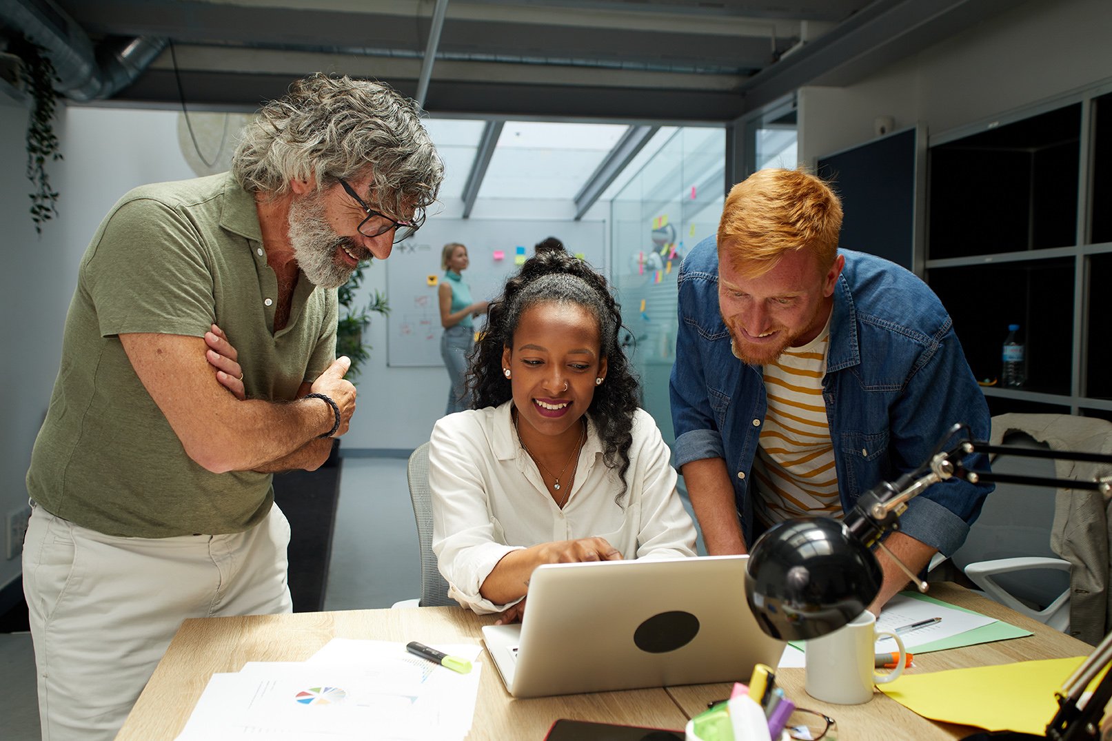 Trois personnes souriantes regardent un ordinateur portable sur un bureau de bureau dans une salle de réunion moderne, avec une femme en arrière-plan écrivant sur un tableau blanc.