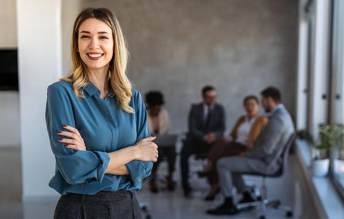 Femme souriante et confiante en premier plan, groupe de personnes en réunion en arrière-plan dans un bureau moderne.